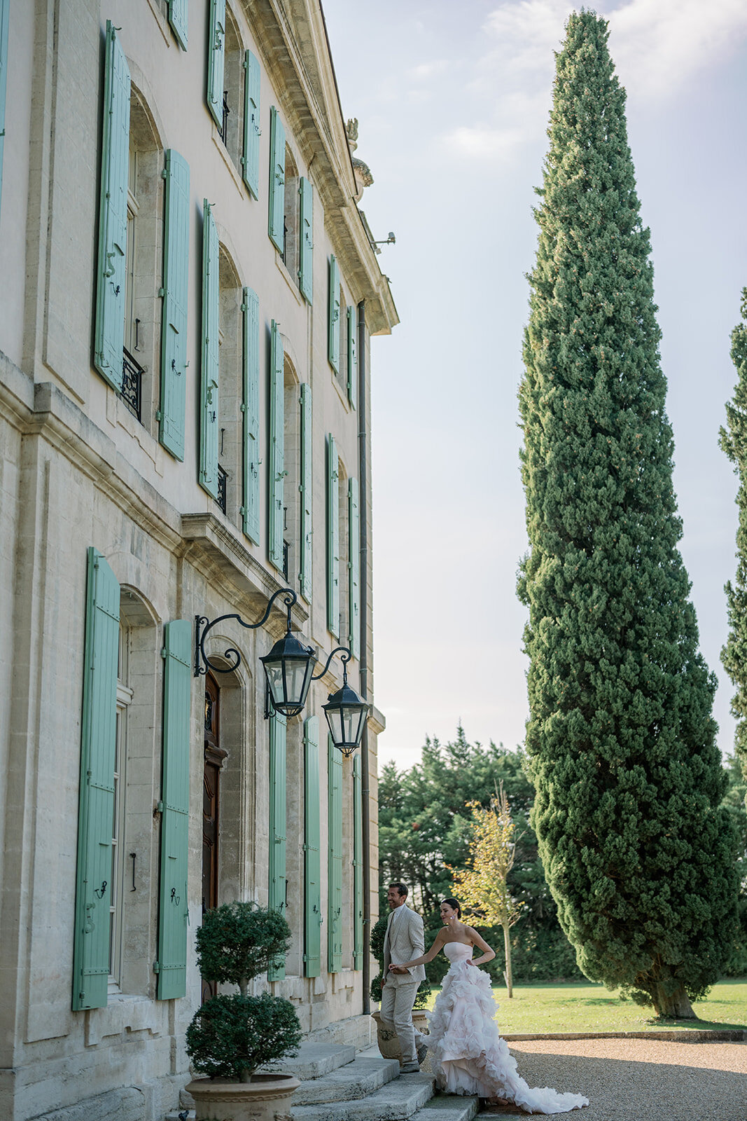 Bride and groom walking beside tall mint-green shutters and stone walls of Château de Tourreau.