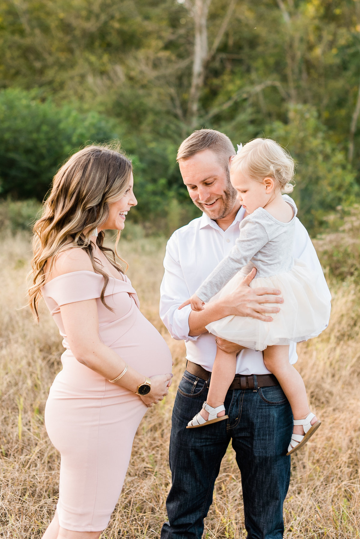 Dad holds toddler while she points at her mom's pregnant belly during a Raleigh maternity photo session. Photographed by Raleigh Maternity Photographer A.J. Dunlap Photography.