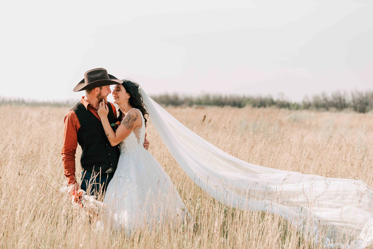 Bride and groom standing close in a pasture with her long veil blowing behind her.