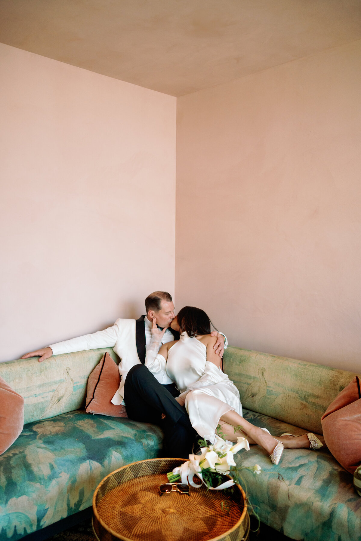 Japna and Chris sharing a romantic kiss on a vintage green sofa against a soft pink wall during their elopement.