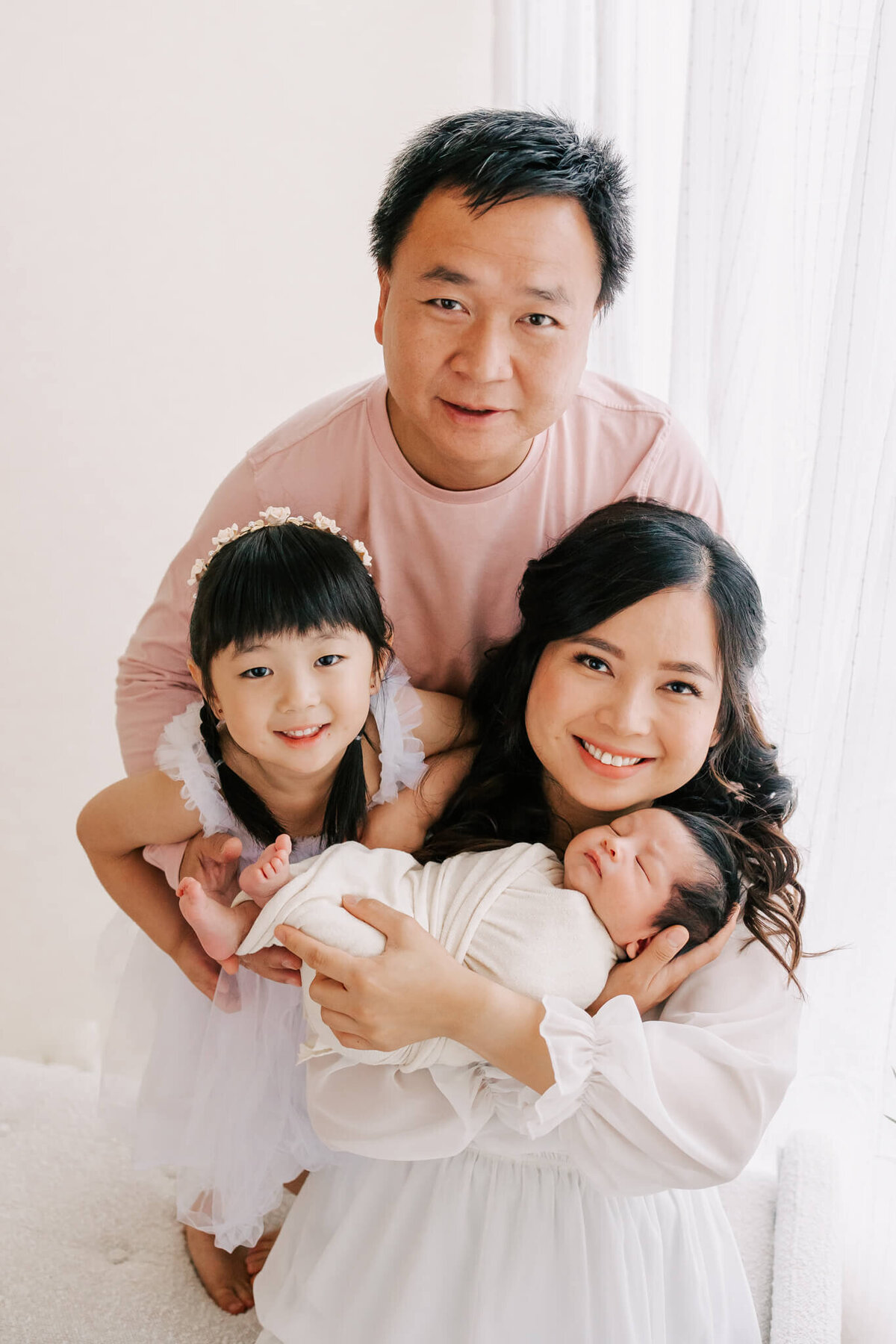 family portrait of a mom, dad, four year old girl, and newborn baby boy. they are all wearing white except for dad who is wearing pink