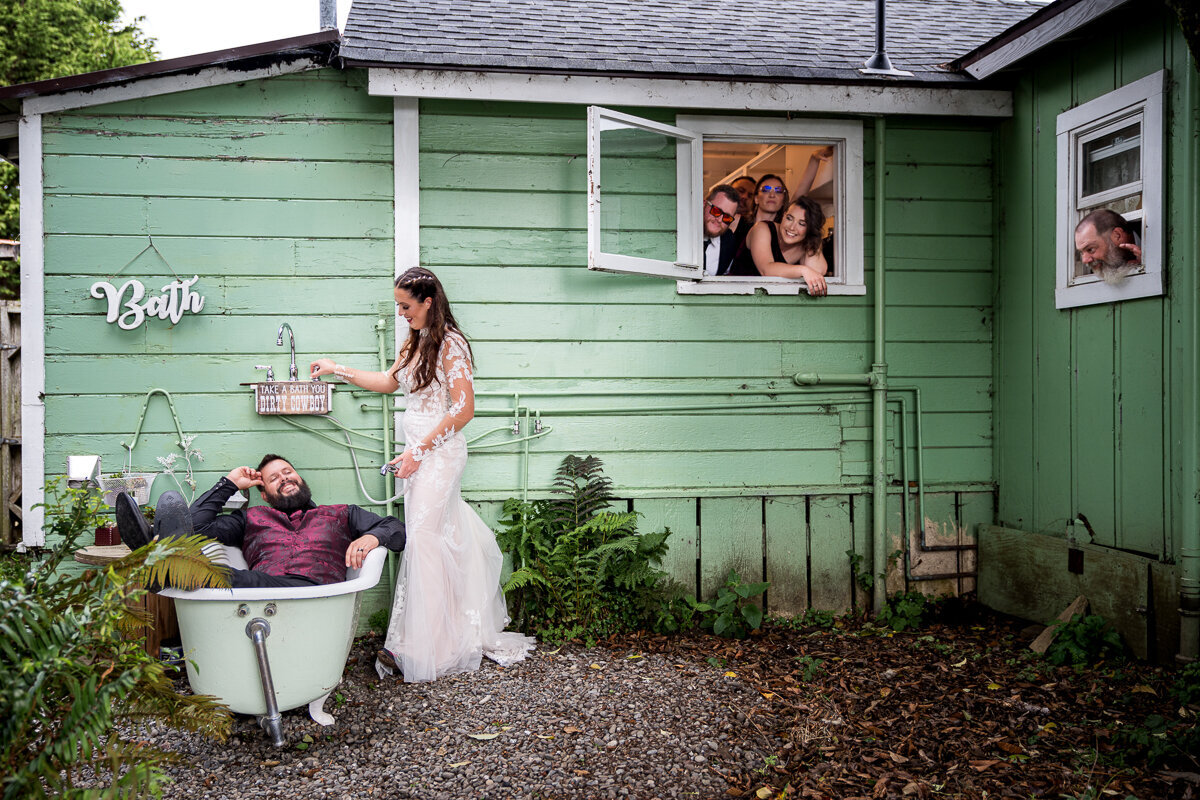 A whimsical and fun wedding photo at North Coast Hitching Post, featuring the groom lounging in an outdoor bathtub while the bride playfully prepares to spray him, as the bridal party peeks through nearby windows, creating a charming Norman Rockwell-style scene.