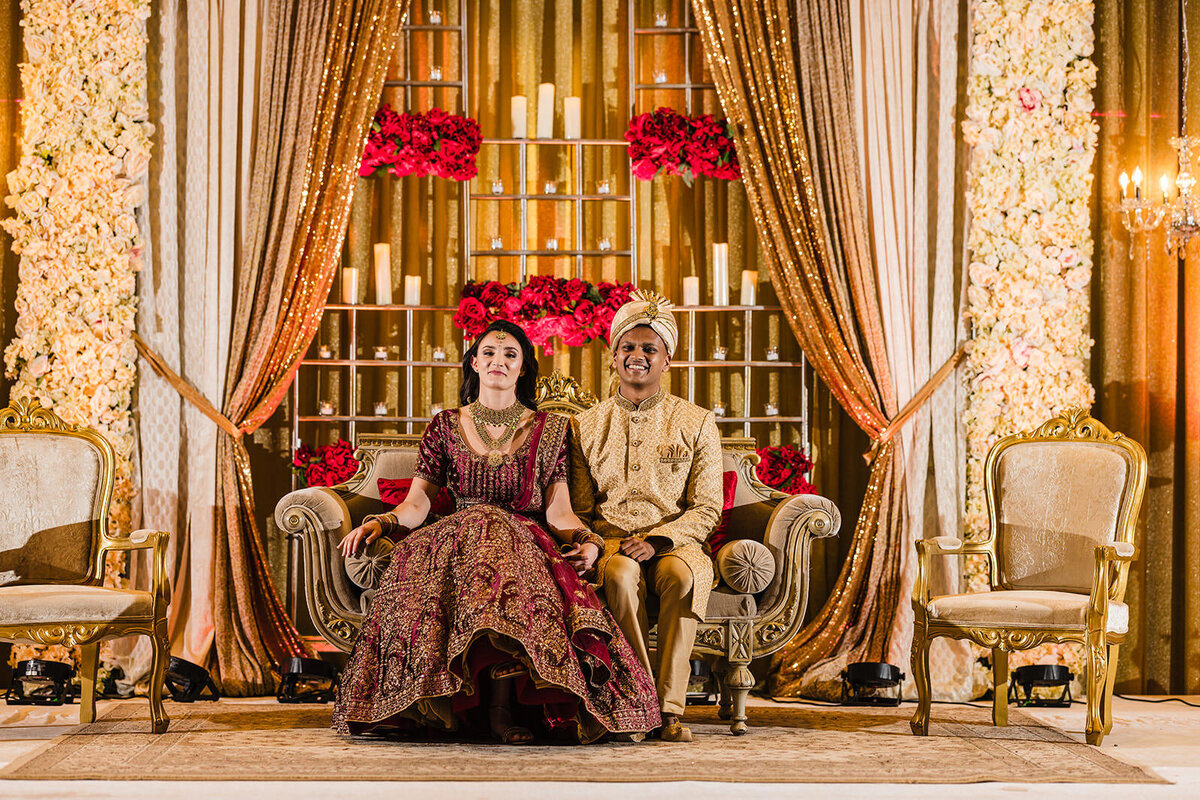 A bride and groom in traditional Indian wedding attire seated on a luxurious sofa on the decorated stage, smiling broadly.