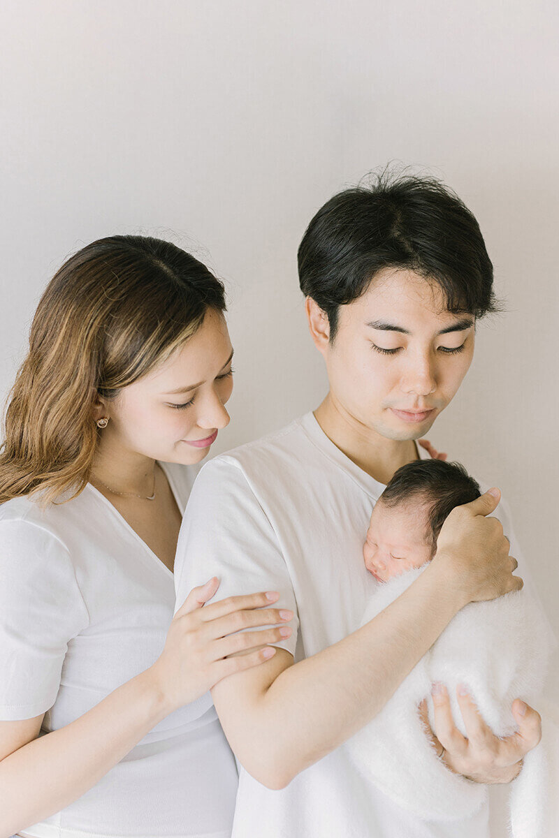 Father is holding newborn baby close to his chest and the mother is standing behind him and smiling to the baby. Newborn photographs shot by Yukika Ishikawa Photography serving Tokyo.