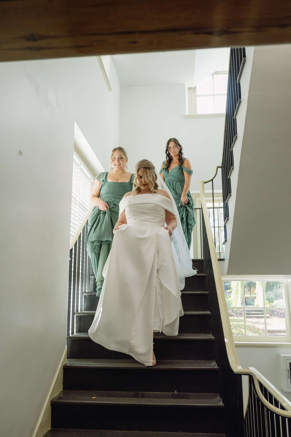 bridesmaids help the bride walk down a stair case from her bridal suite with the bridesmaids in sage green bridesmaids dresses holding the brides train