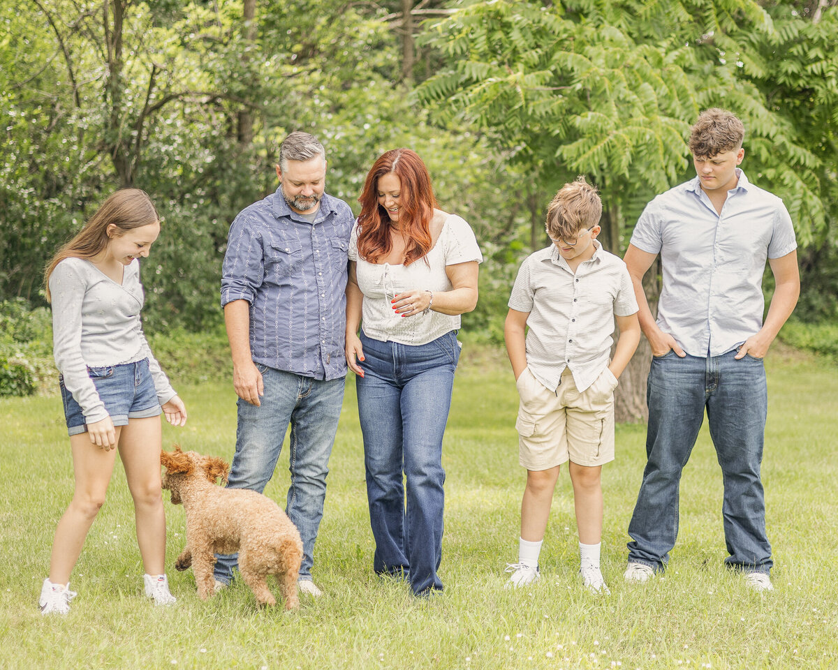 Family of five and their brown dog standing on grass with a tree line behind them.  Everyone is looking down at their dog with some smiling and some not.  Dog is trying to walk away and is facing away from the camera.  Daughter is wearing blue jean shorts, a long sleeve light blue shirt, white shoes, and has long light brown hair.  Dad is wearing blue jeans, blue and white stripped button up shirt, and has short pepper hair and beard.  Mom has long red hair, wearing a white short sleeve shirt and blue jeans.  Younger son is wearing a light tan button up and khaki shorts, white shoes, short light brown hair.  Oldest son is wearing a light blue short sleeve button up, blue jeans, and has short light brown hair