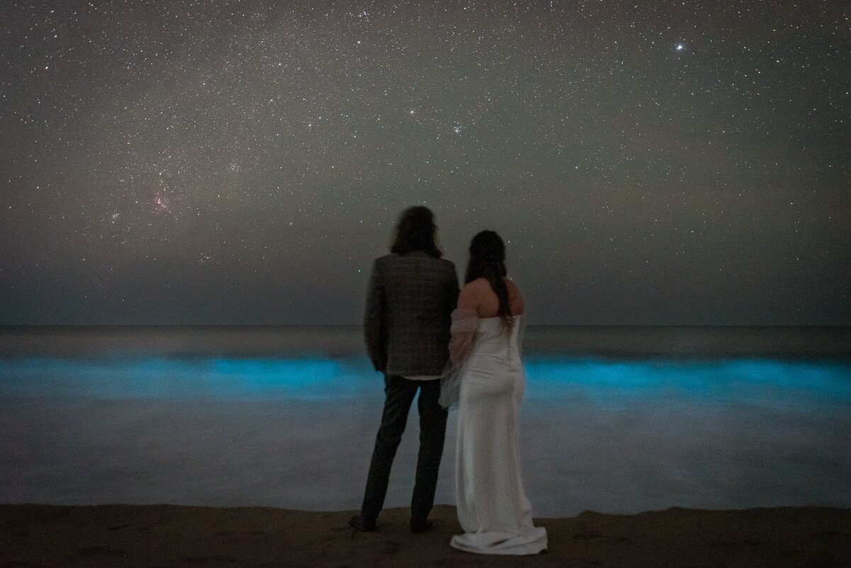A bride and groom in their wedding finest stand in the dark night during their adventurous luxury Oaxaca Mexico Elopement.  The night sky is littered with stars and in front of them filling the bottom half of the frame, a bright blue and milky white bioluminescent wave crashes ashore. 