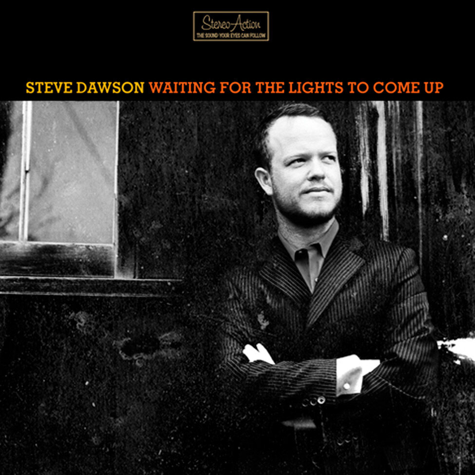 Album Cover Steve Dawson Title Waiting For The Lights To Come Up Artist Steve Dawson standing with arms crossed against his body wearing a black suit grey shirt and tie by black building with window black and white