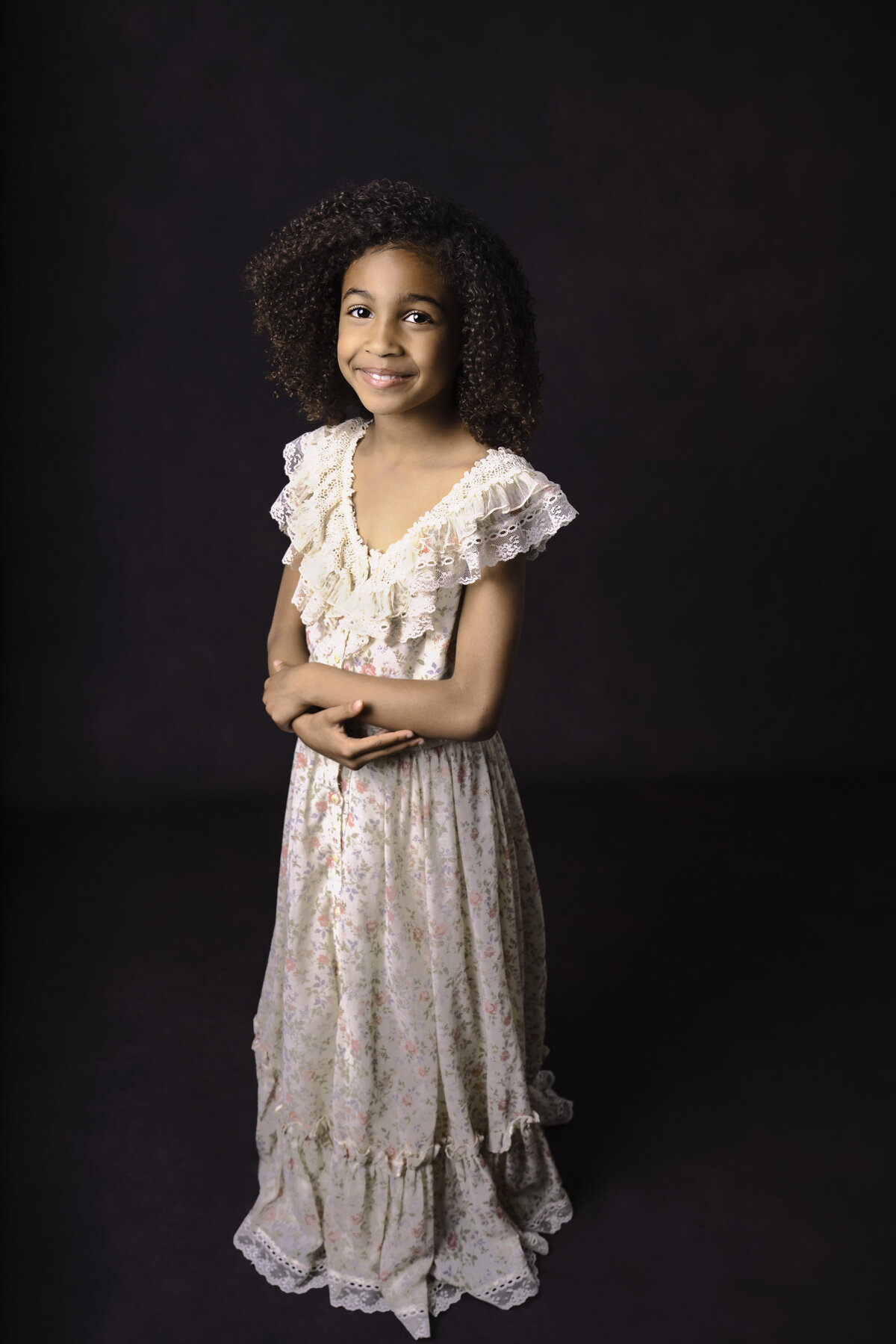 Little girl in floral dress poses for portrait. Her arms are folded gently at her waist and she is looking at the camera smiling. Captured by Liza Nicole Photography, Philadelphia and Main Line family photographer.