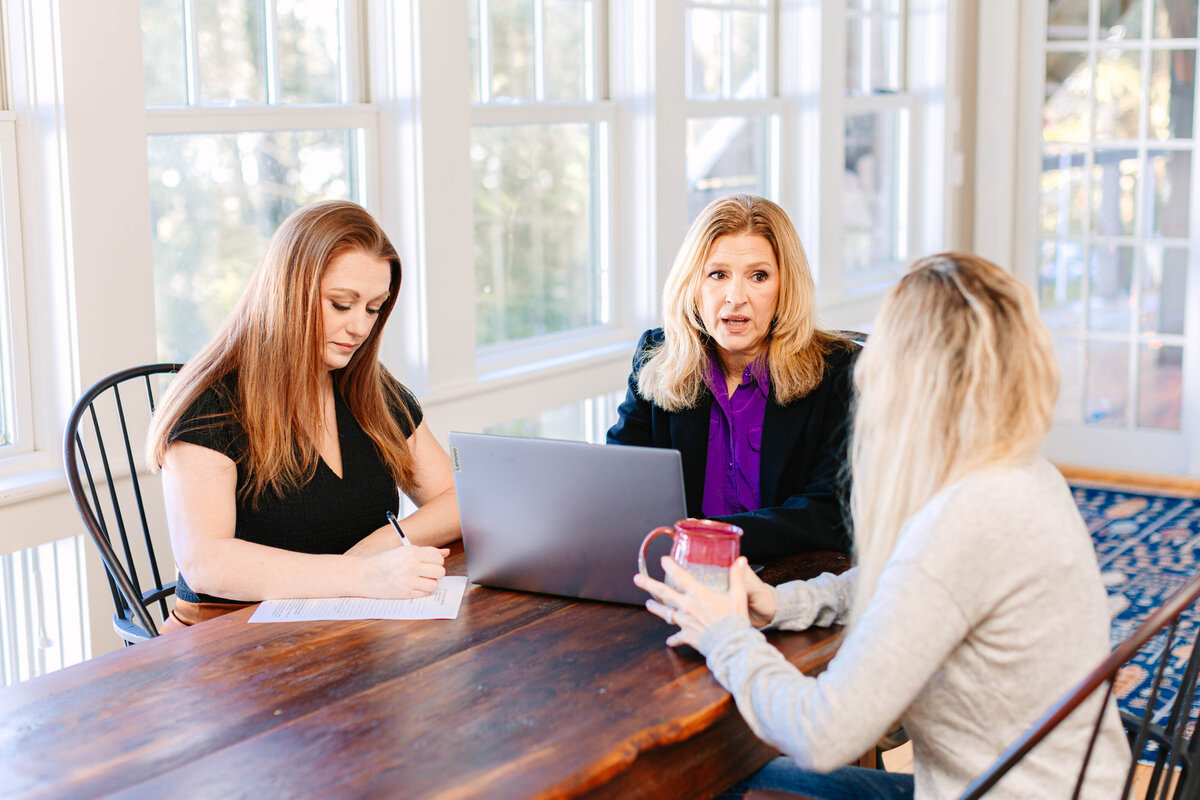 Three women have a discussion at a wooden table in a bright, modern space. One woman in a black blazer speaks while another in a black top takes notes. The third woman, with long blonde hair, listens while holding a red mug. A laptop and documents are spread across the table