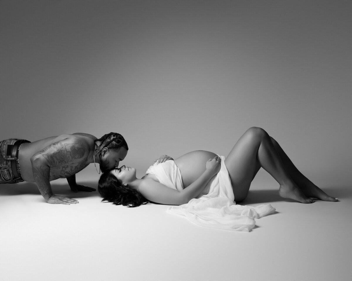 Intimate studio maternity portrait of a couple in black and white, with the partner kissing the expecting mom's forehead.