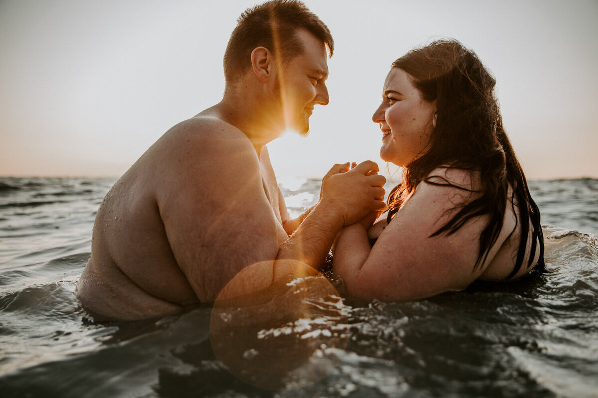 Man and woman sitting in Lake Huron at Grand Bend Beach in waist-deep water. They are facing each other and he is clasping her hands under her chin. The sun is setting behind them creating golden light and sun flare.