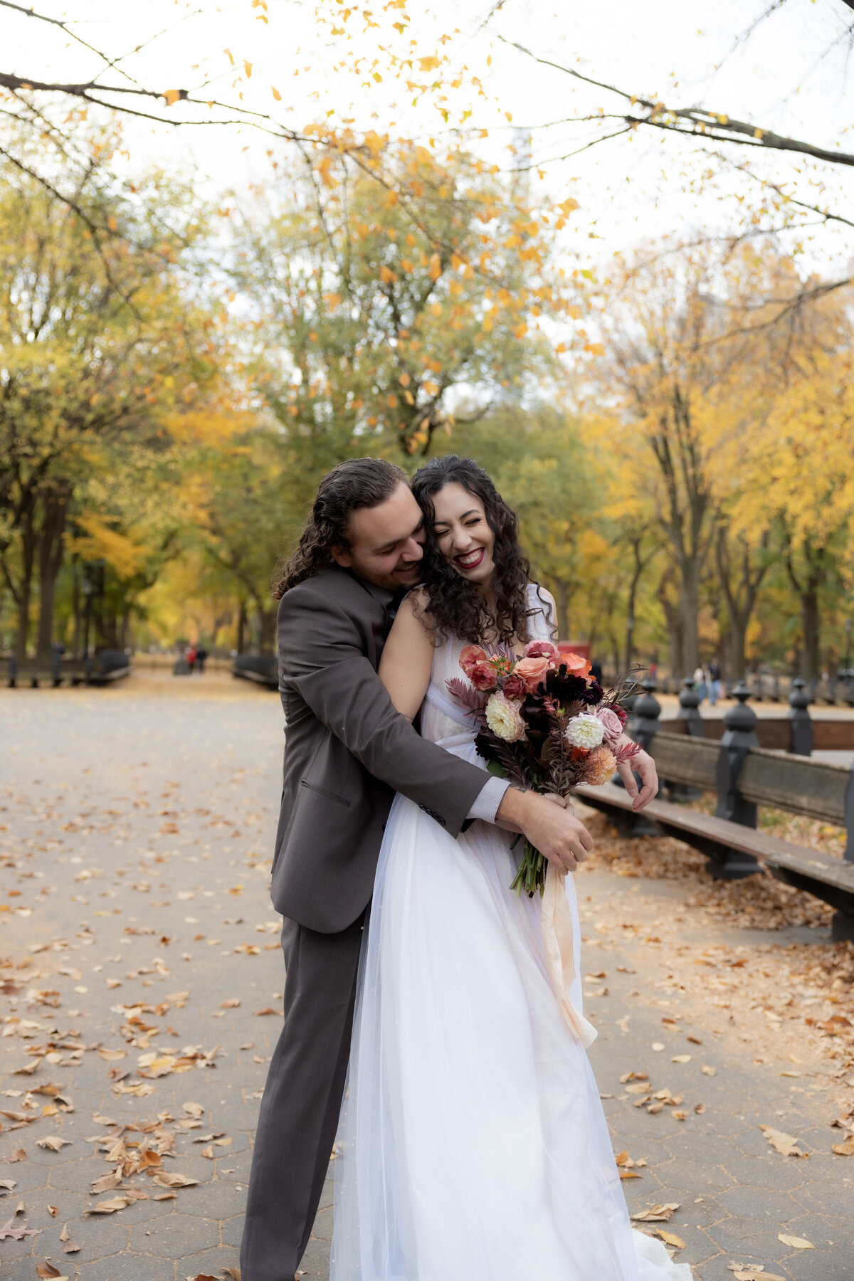 The bride laughing joyfully while holding her bouquet among autumn leaves in Central Park.