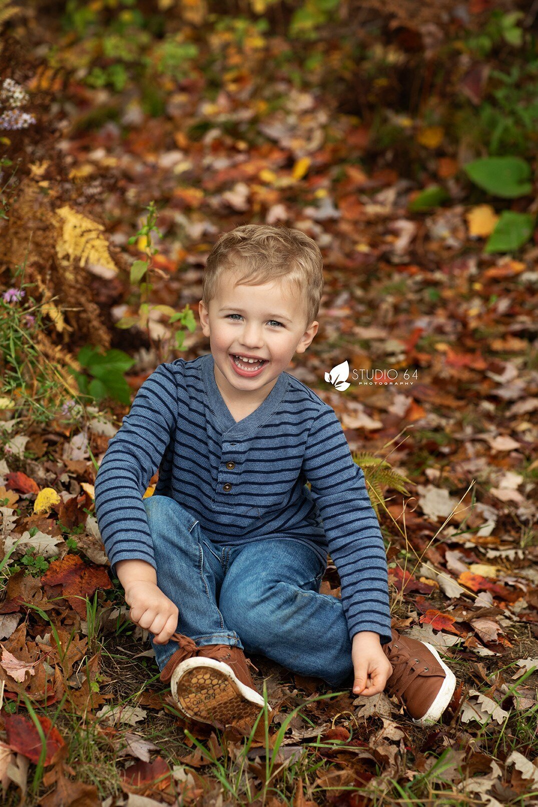 A five year old boy sitting on a ground full of leaves being silly and smiling.