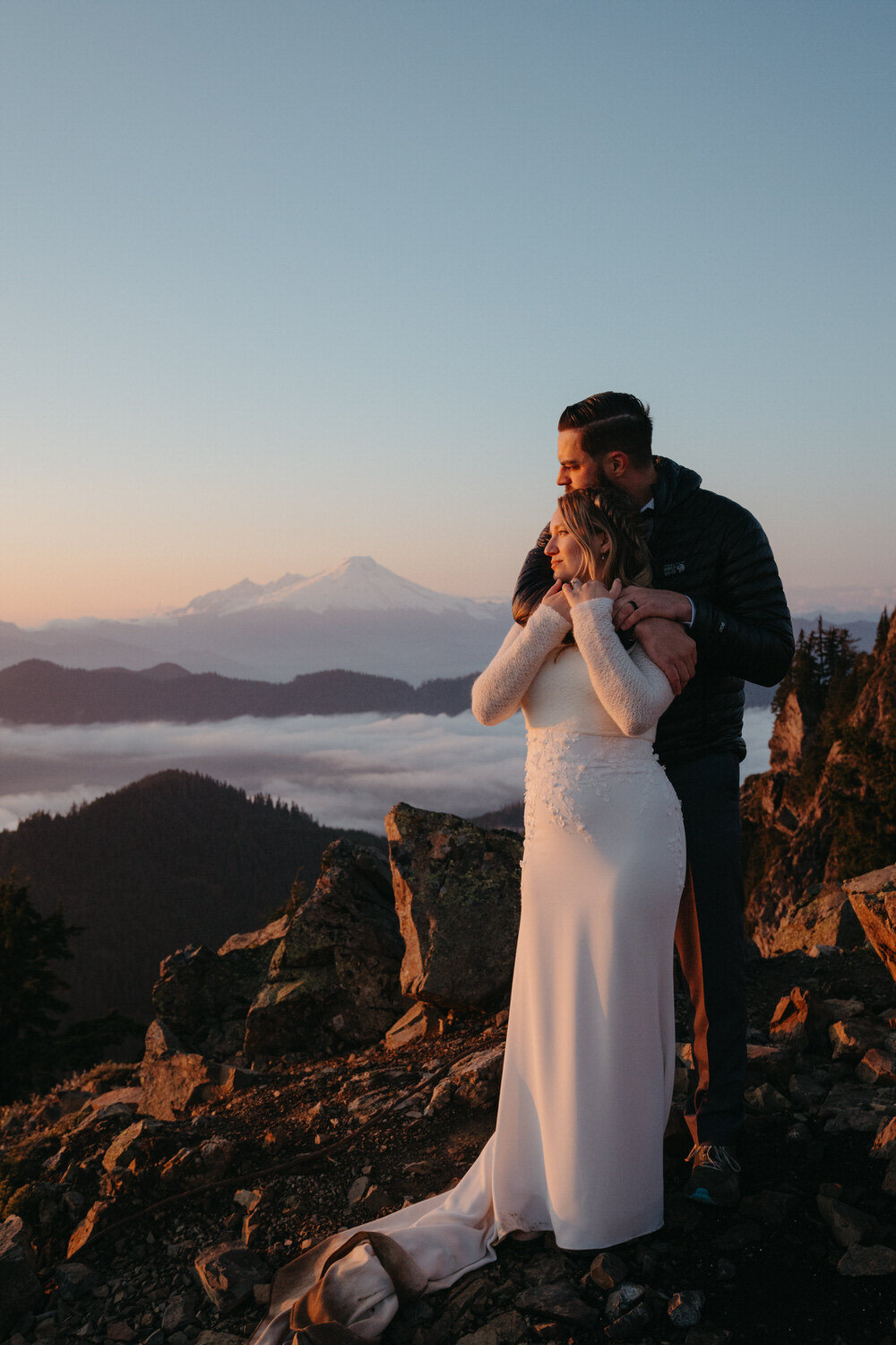 Hiking elopement on top of a mountain, groom hugs bride as she looks at the sunset