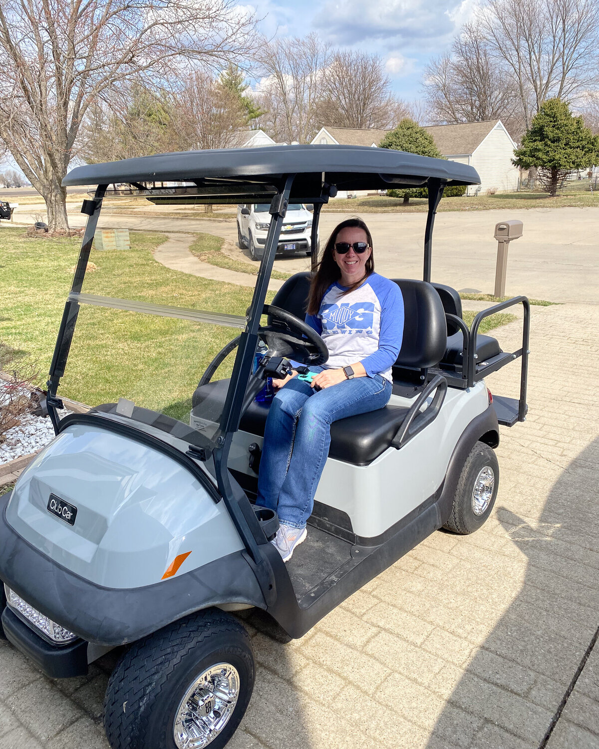 woman sitting on a gray golf cart in her driveway, smiling at the camera, wearing sunglasses