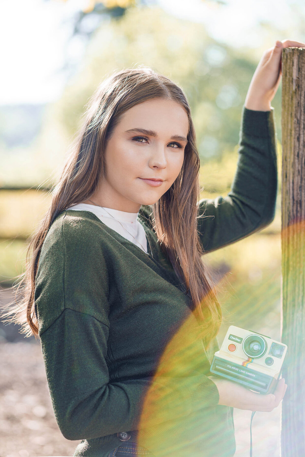 Senior Graduate girl teen in green sweater on farm leans on post with camera in hand and sun flare adds dreamy feeling