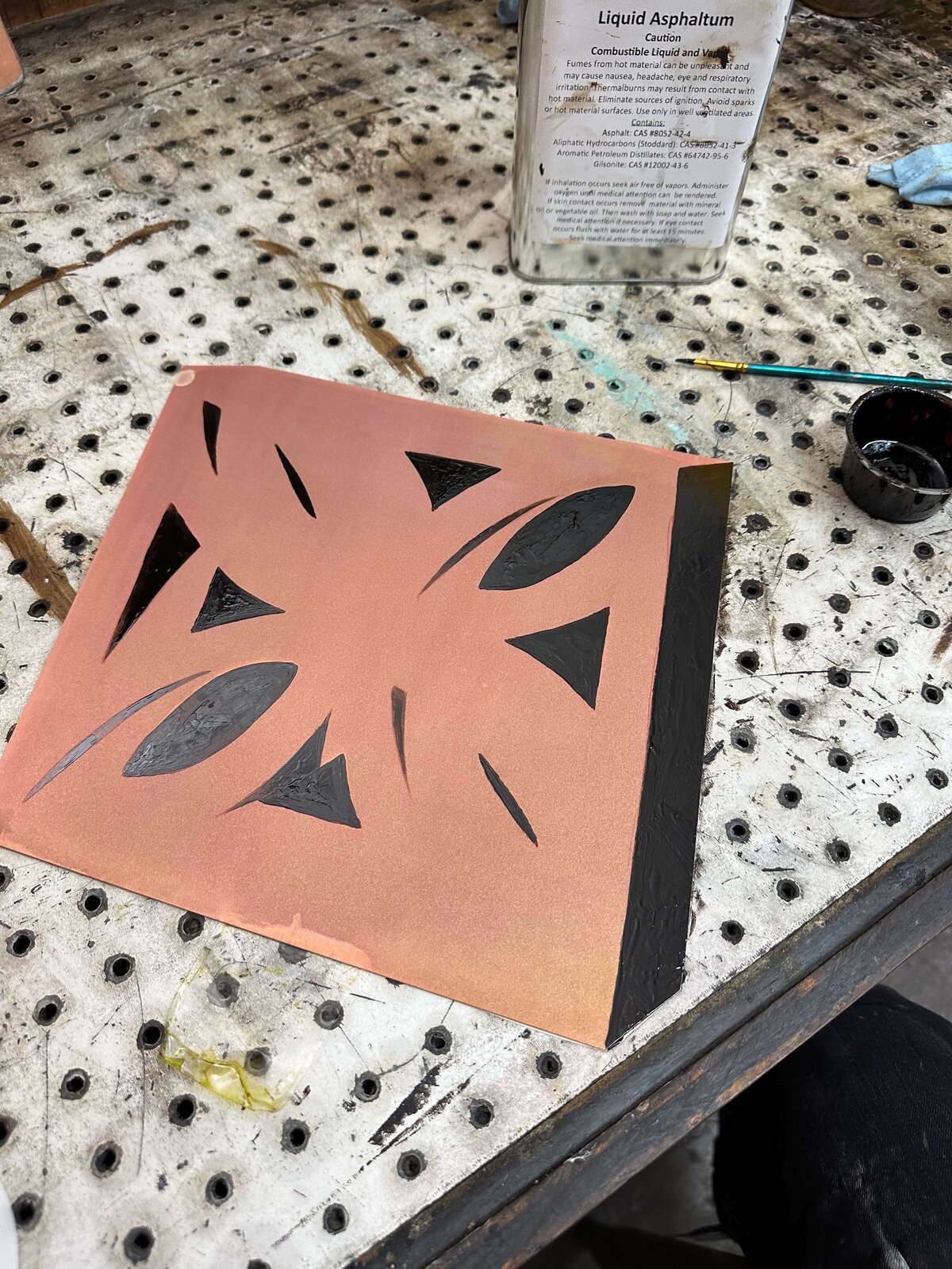 A copper plate with abstract black asphaltum-painted shapes, ready for aquatint etching, sitting on a perforated metal surface.