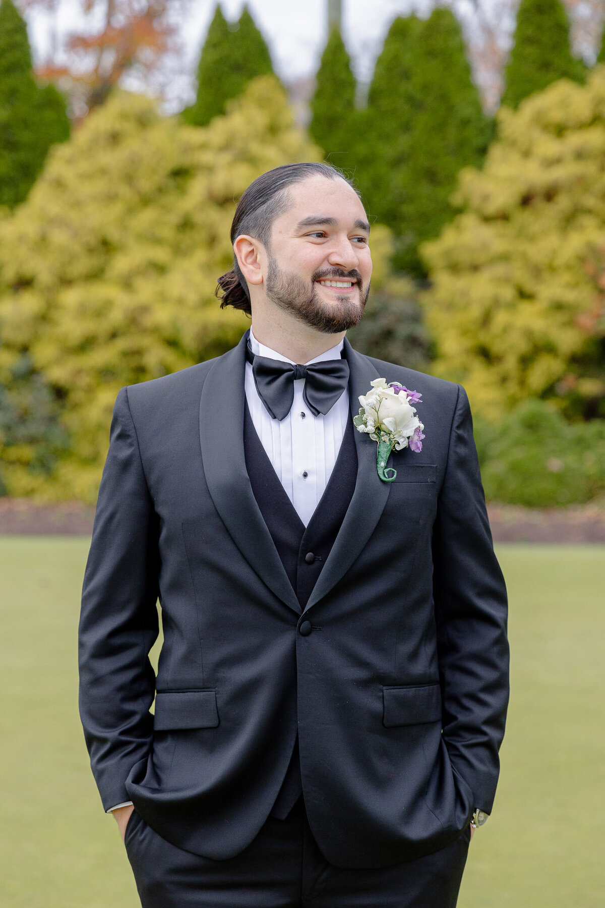 A groom smiles on his wedding day. His hands are in his pockets and he's looking off to the side as he's standing outside.