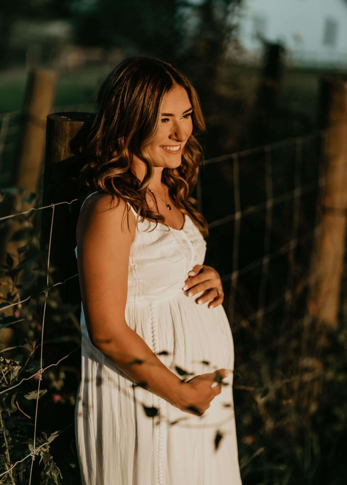 A pregnant woman in a white dress leans against a fence, beautifully captured by a Pittsburgh maternity photographer.