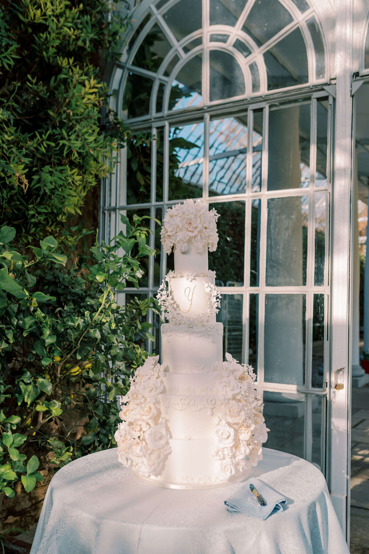 five tier white luxury wedding cake with elaborate white sugar flowers with green foliage in the background in the orangery at avington park