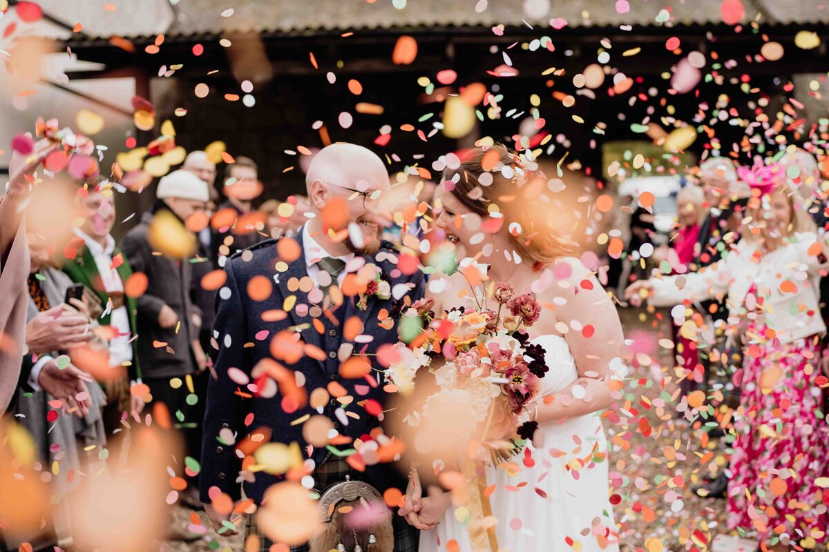 A couple strolls hand in hand through a shower of colourful confetti in the courtyard of a farm, beaming with joy. The bride, photographed in her white wedding dress and bouquet, and the groom, sharply dressed in his suit, are surrounded by joyful guests. The Aberdeen sky is cloudy above them.