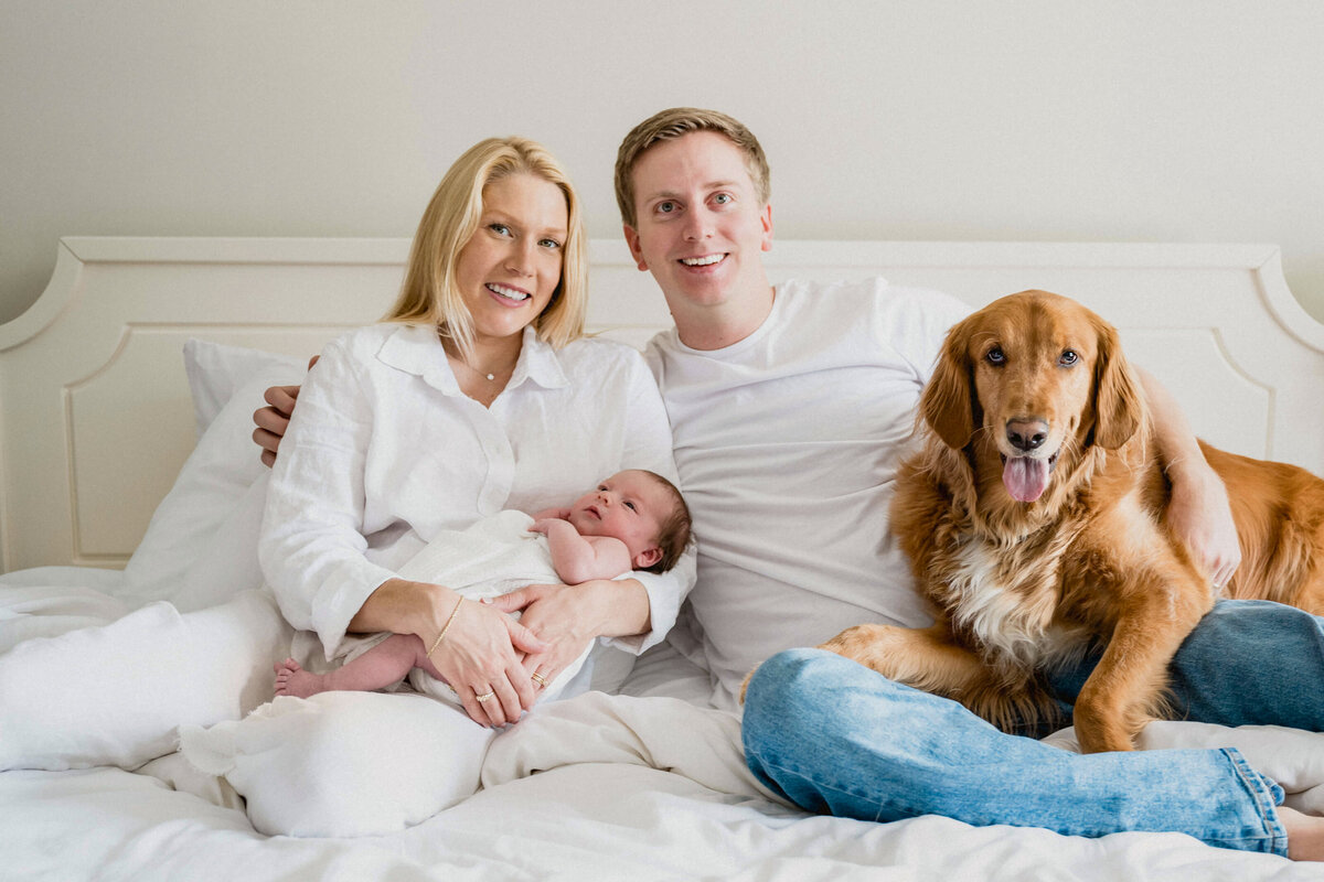 Family with 5 day old baby and Golden Retriever on bed looking at camera.