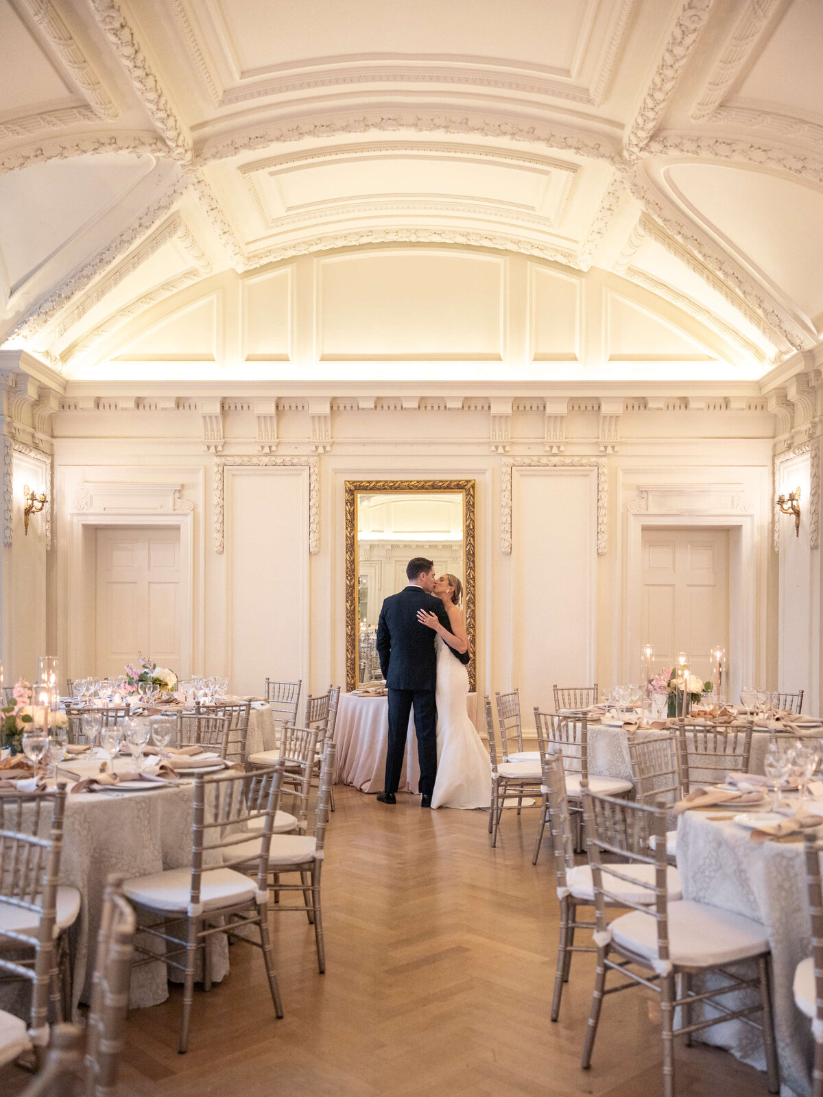 A couple, dressed in wedding attire, embrace in an elegant ballroom with ornate ceilings. The room features round tables with floral centerpieces and candlelit settings, all in a soft, romantic ambiance.