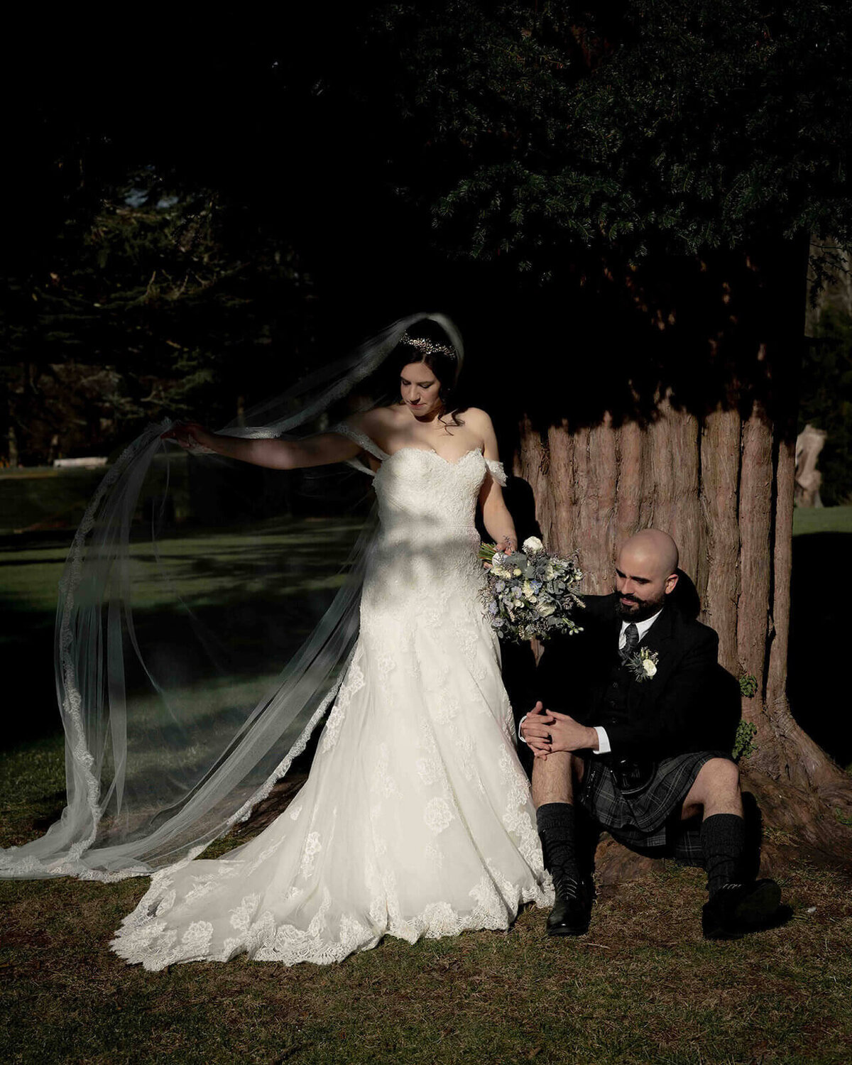 Wedding portrait with groom sat at a tree stump with his bride elevated in a standing position playing with her veil in the wind.