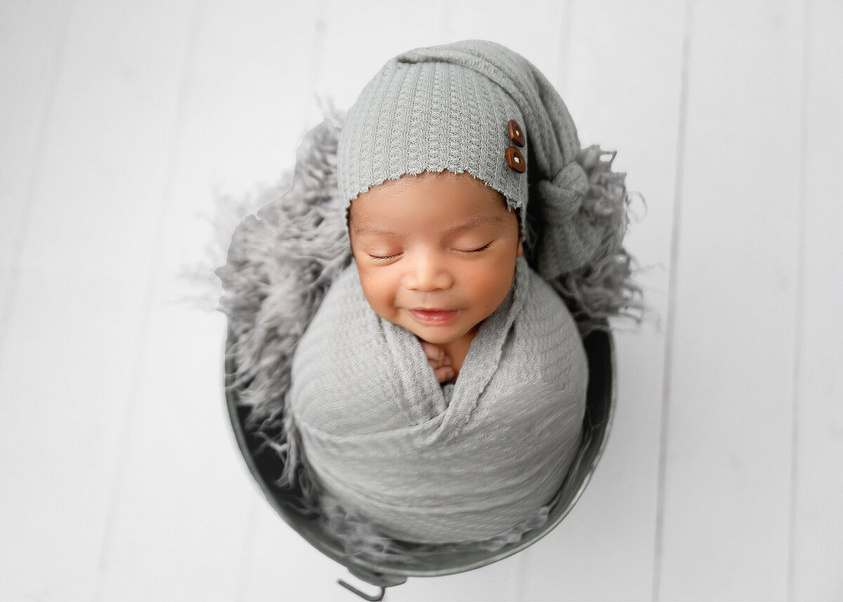 Smiling baby boy swaddled in gray wrap wearing a sleepy hat posed in a bucket