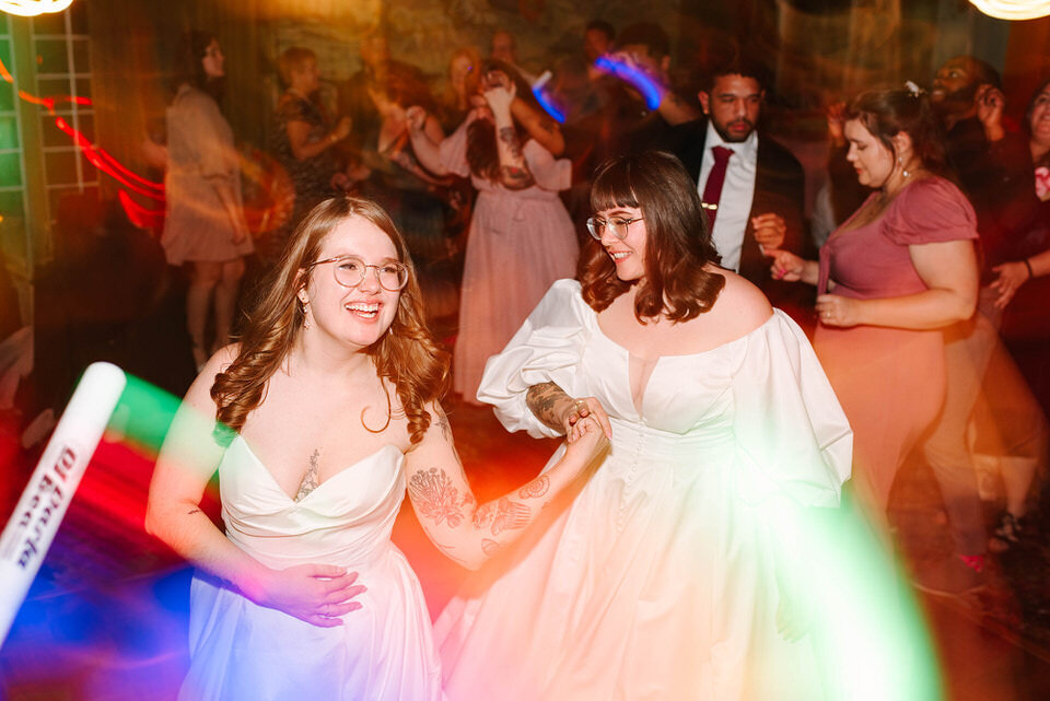 Two brides holding hands and laughing on the dance floor at their wedding, surrounded by friends and colorful motion blur lights, captured in a joyful, candid moment.