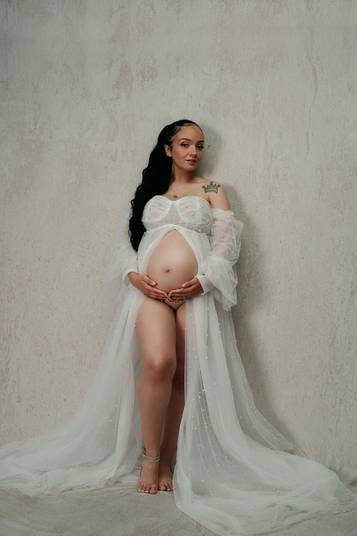 Pregnancy portrait of woman leaning against white backdrop wearing a long white dress open to see belly