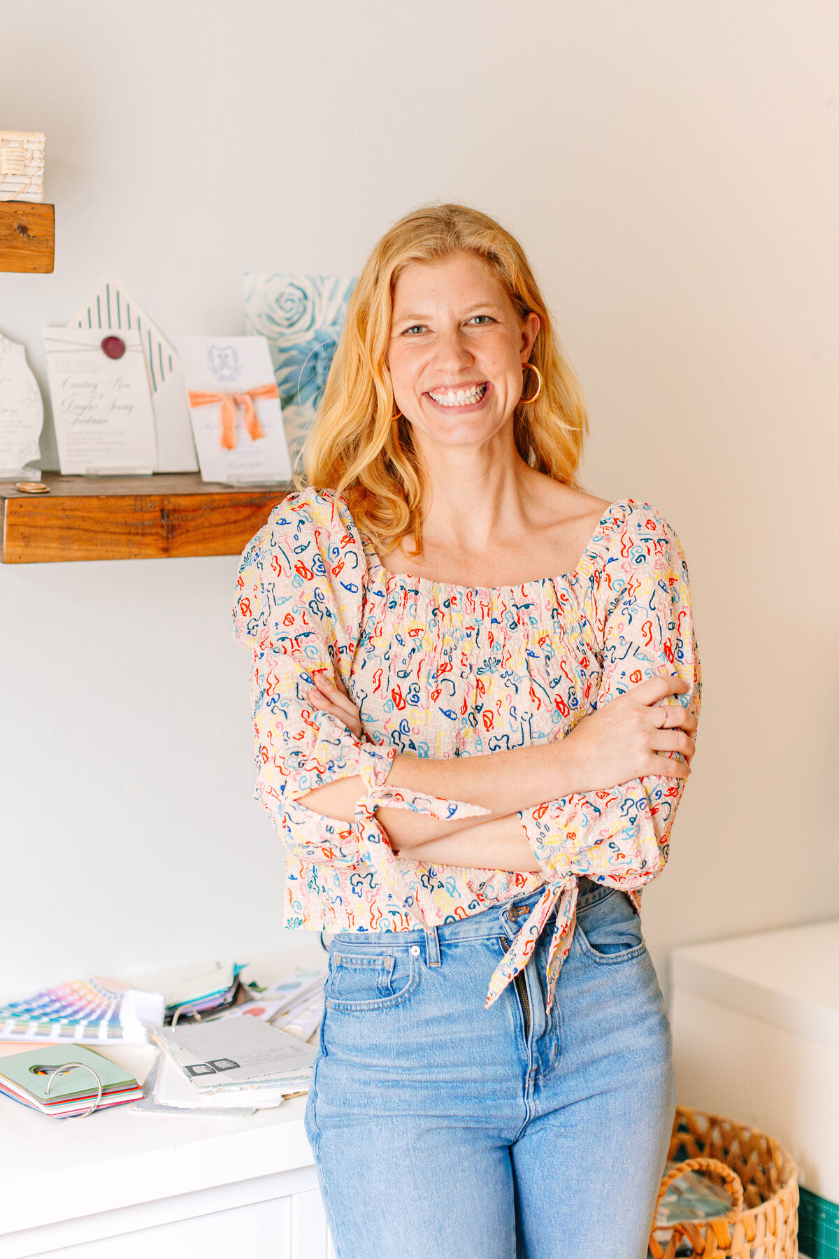 A smiling woman with wavy blonde hair and gold hoop earrings stands with her arms crossed in a creative workspace. She wears a colorful patterned blouse and jeans. Behind her, a wooden shelf holds stationery samples, and a desk is covered with color swatches and design materials.