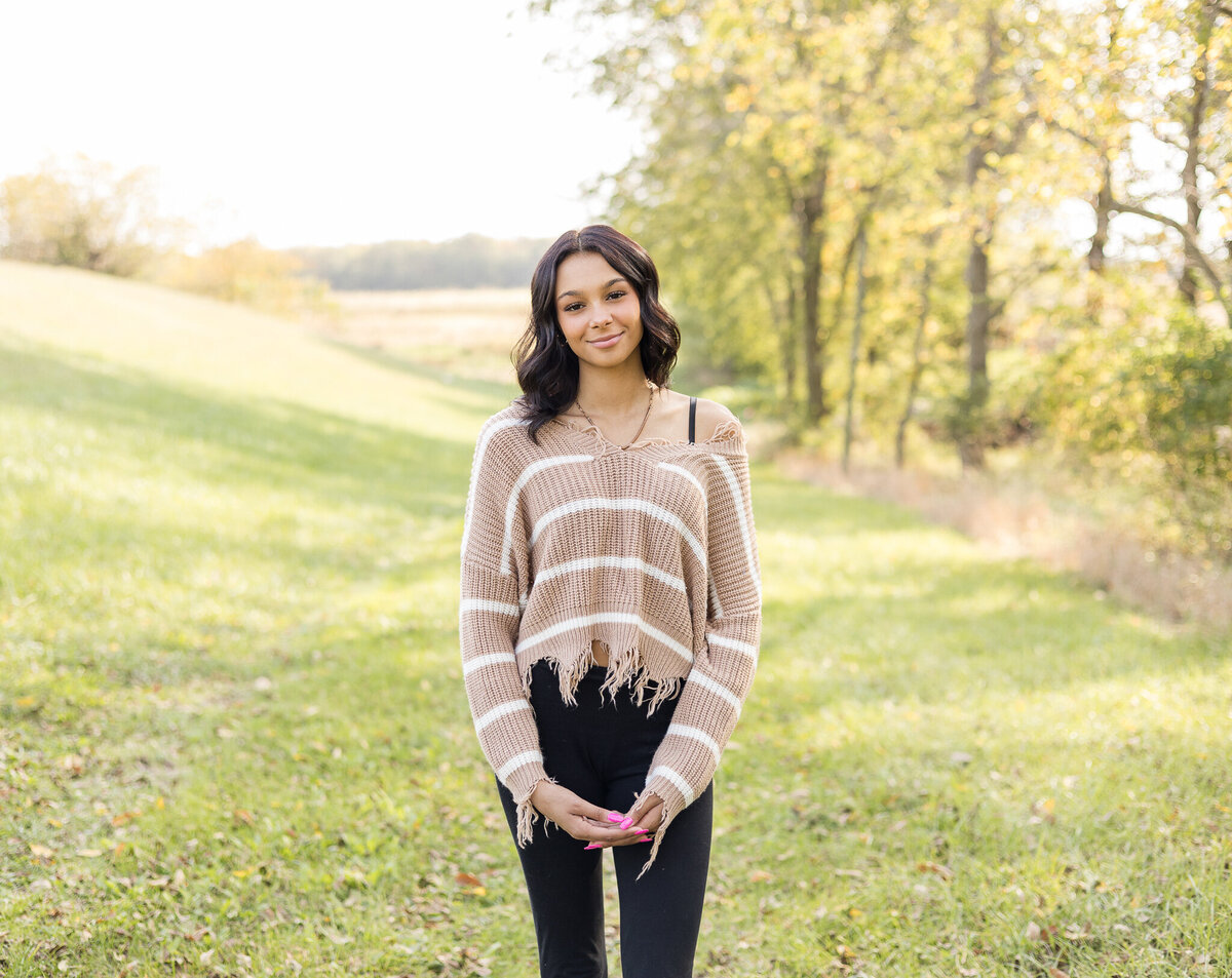 high school female senior, with long black hair, standing in grass with a tree line next to her, looking at the camera smiling softly, wearing black leggings, and light tan and white stripped shirt