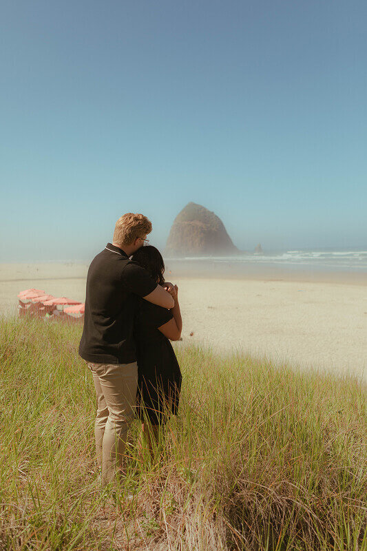 A newly engaged couple look off into the distance at Haystack rock during their engagement proposal photos.