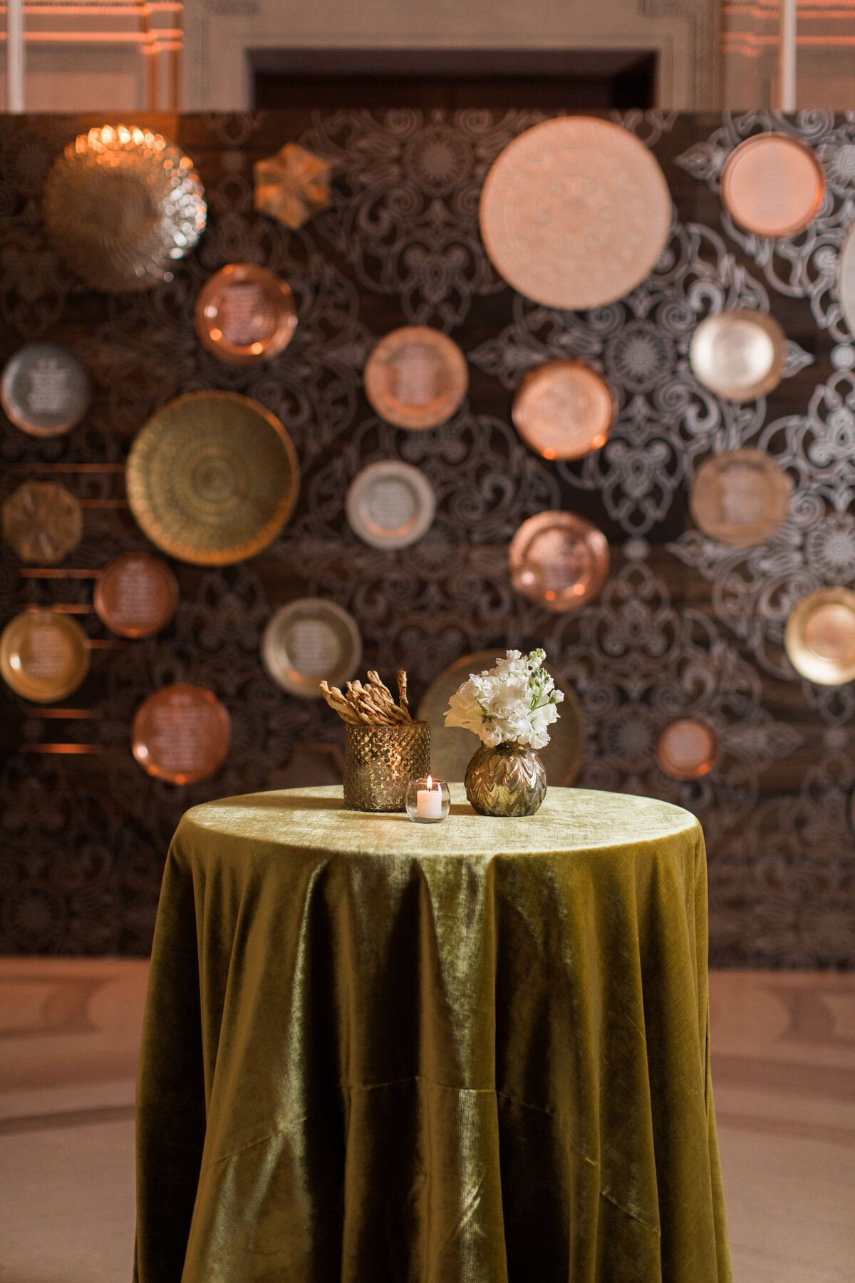 The photo was taken at the Mellon Auditorium in Washington D.C. by photographer Sarah Bradshaw. This elegant image showcases a round table adorned with an olive-green velvet cloth, embellished with a small vase of white flowers, a glowing candle, and a stylish metal container holding pens or pencils. The backdrop of the photo captures an ornate wall adorned with various metallic plates, illuminated by a warm, ambient light. This setting exemplifies the luxurious European flair of the venue, making it a perfect choice for sophisticated events and gatherings.