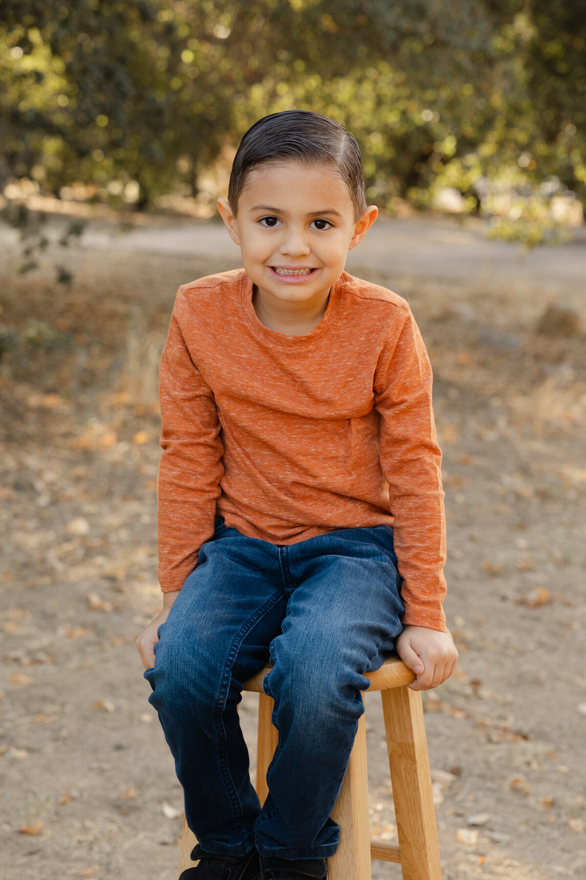 Young boy standing outdoors in orange sweater and jeans, smiling at the camera. Photographed by Yucaipa family photographer Kaitlyn Dawn Photography.