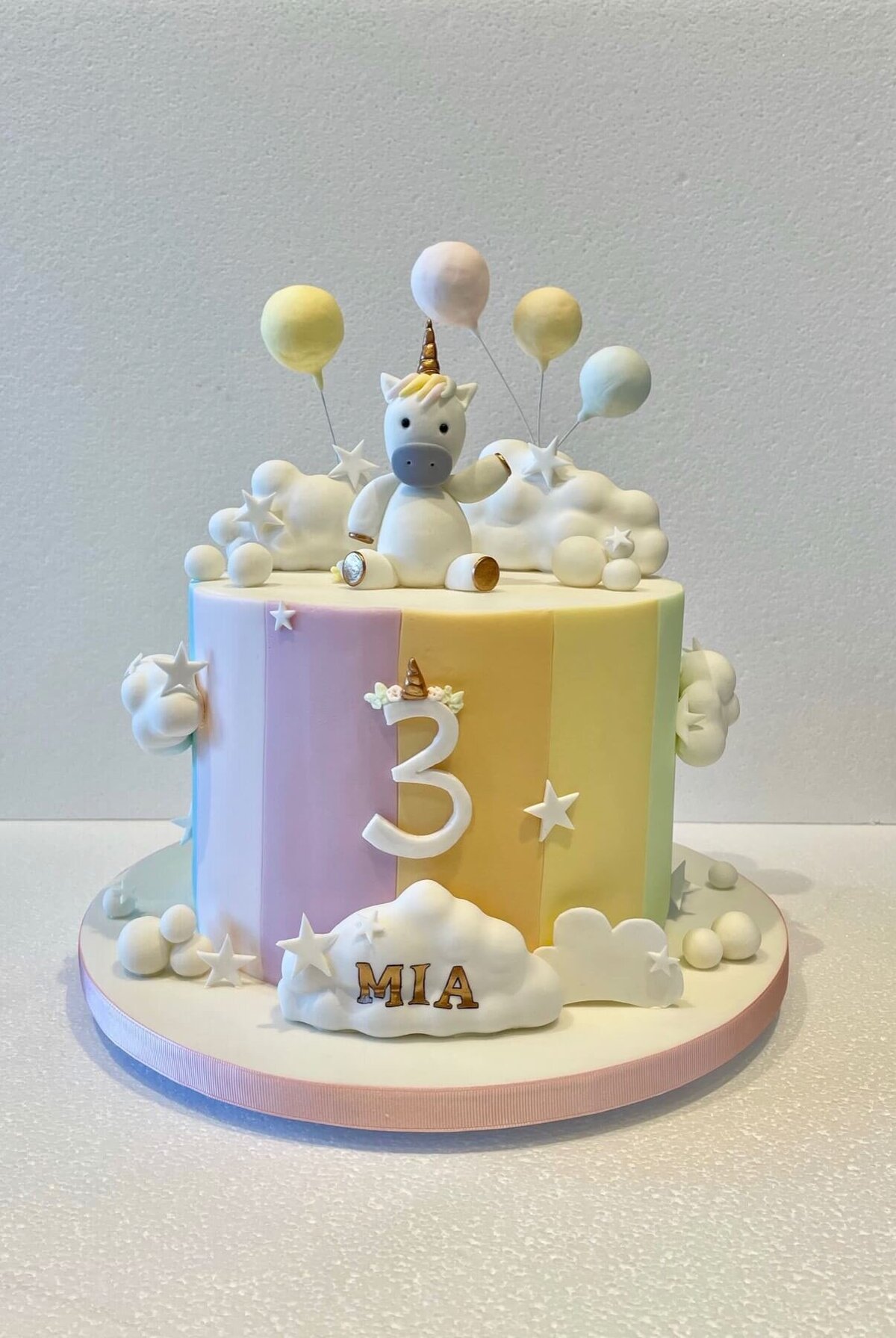 A third birthday cake with a rainbow and clouds theme and a unicorn on top with a party hat and 4 balloons