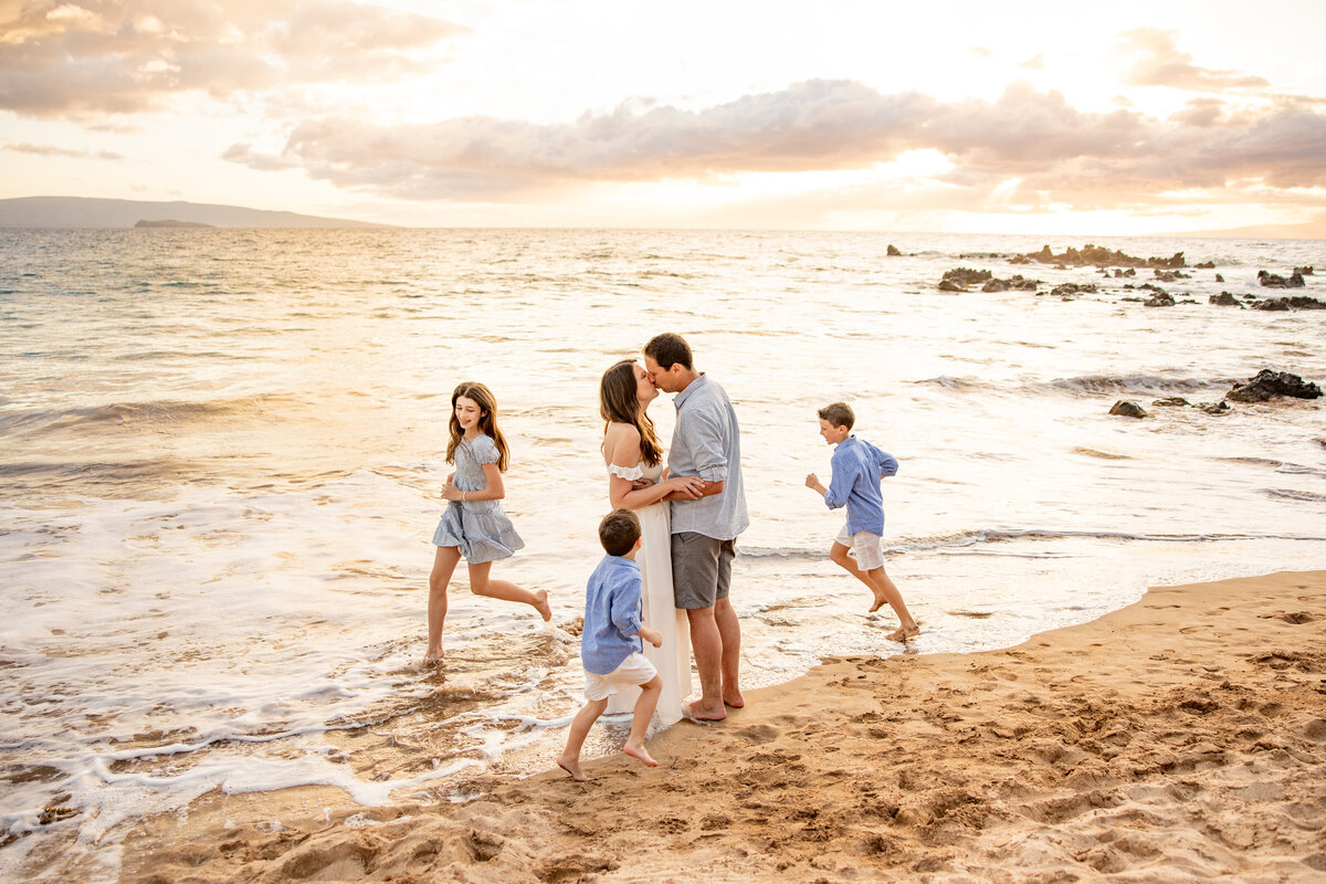 Maui Family Photographers capture mother and father kissing while children run around during family beach photos