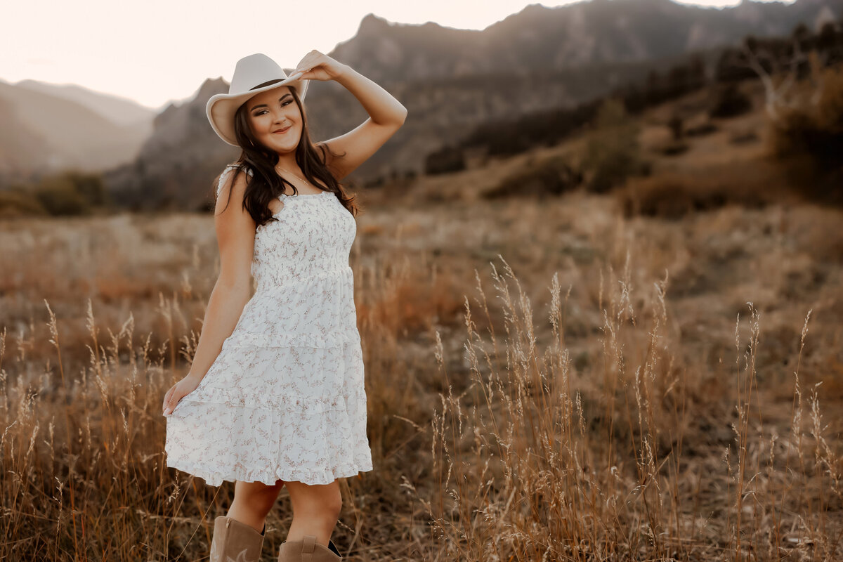 Senior girl in white short dress with white cowboy hat in a golden tall grassy field in Boulder, Colorado