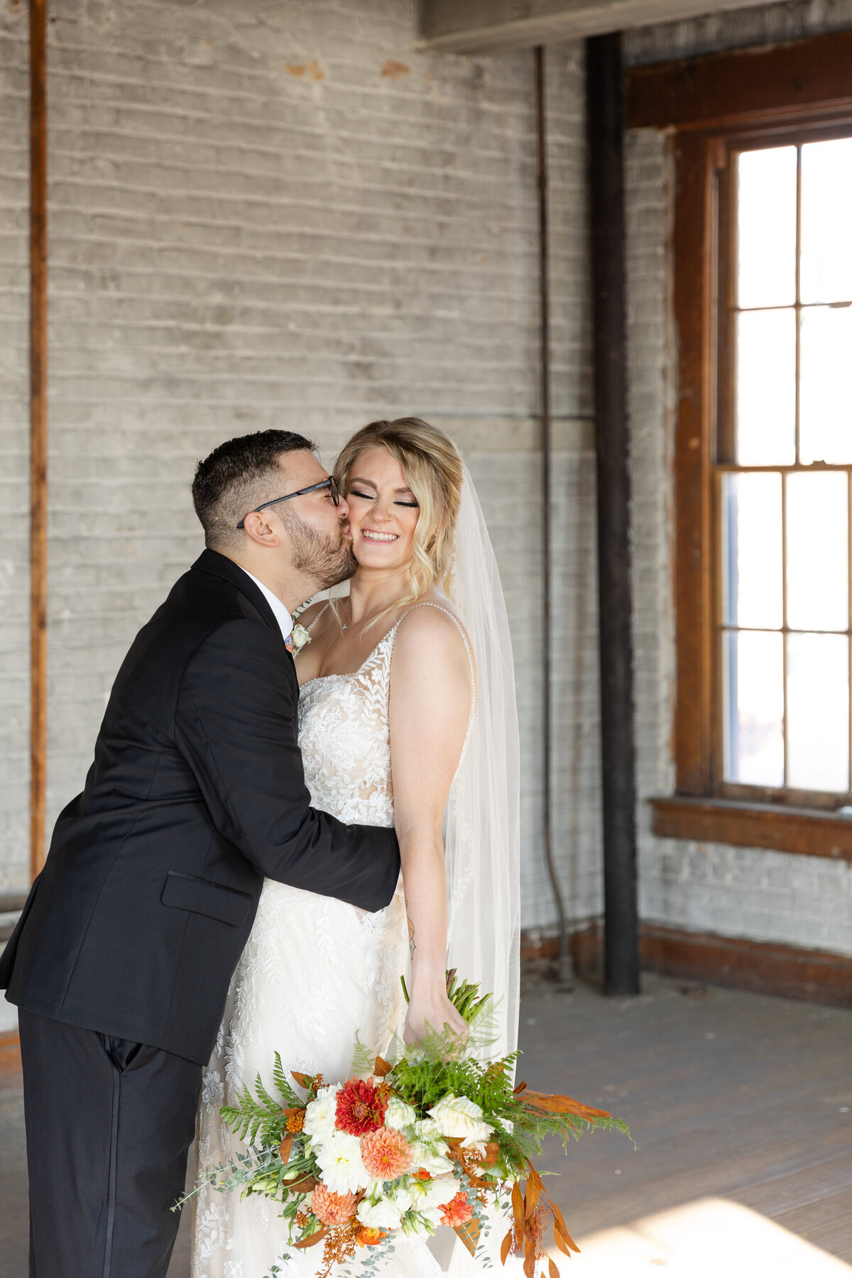 Groom kisses bride's cheek while she holds her bouquet at. her side