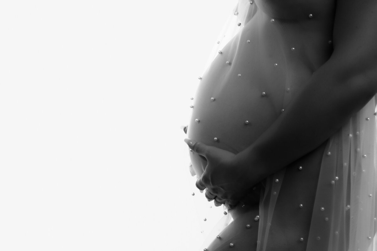 Black-and-white close-up photograph of a pregnant woman gently cradling her belly. She is draped in a sheer, pearl-embellished fabric that adds an elegant, ethereal feel to the image.