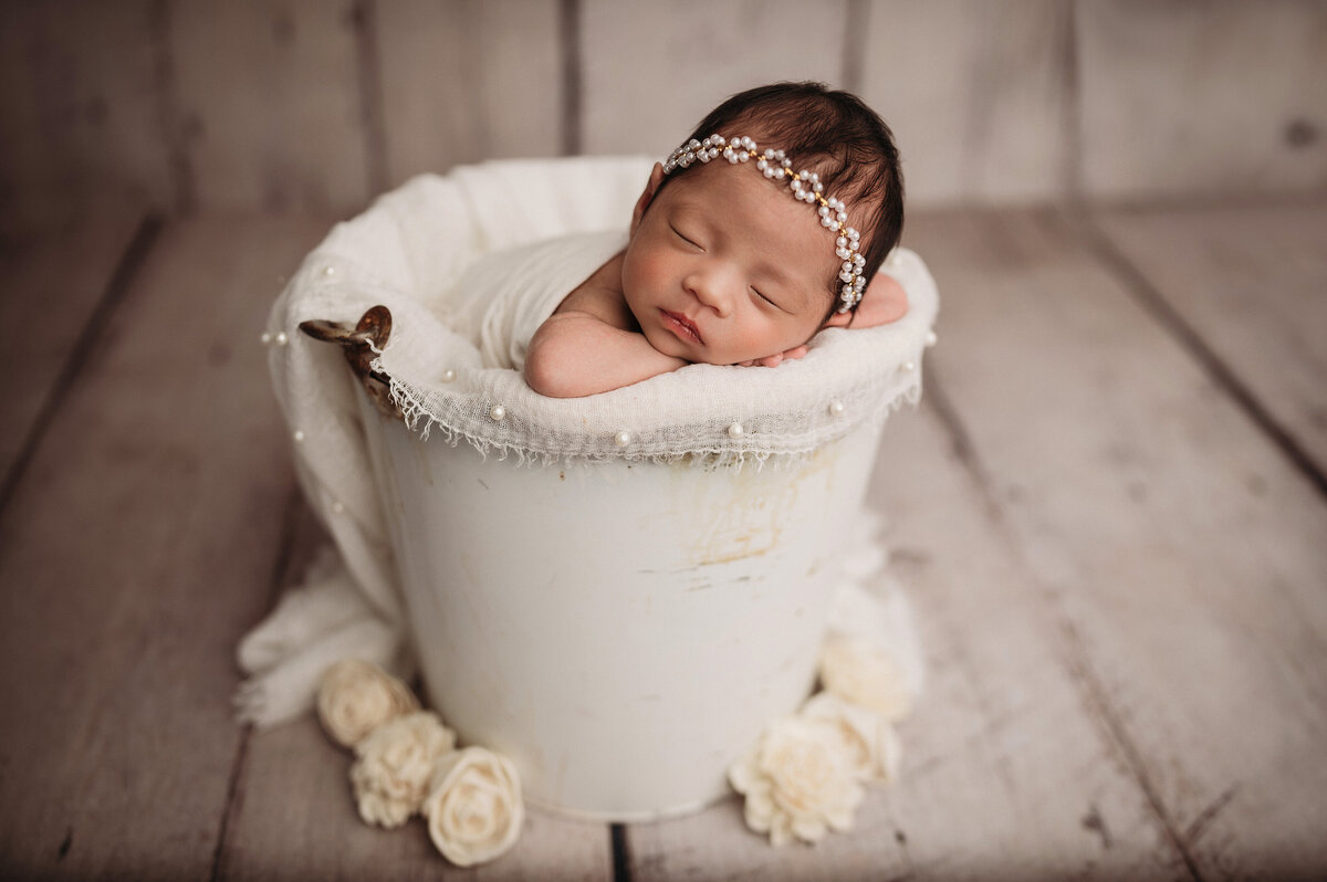 sleeping newborn baby girl  wearing a white floral bow  posed with chin under hands  in a white bucket with pearl white fringe layer on a white wooden floor with white flowers surrounding the bucket