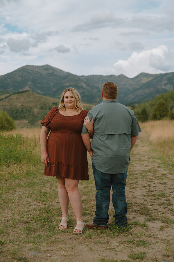 A woman holds the arm of her fiancé and smiles at the camera. Her fiancé is facing away from the camera looking at gorgeous mountains in Jackson WY.