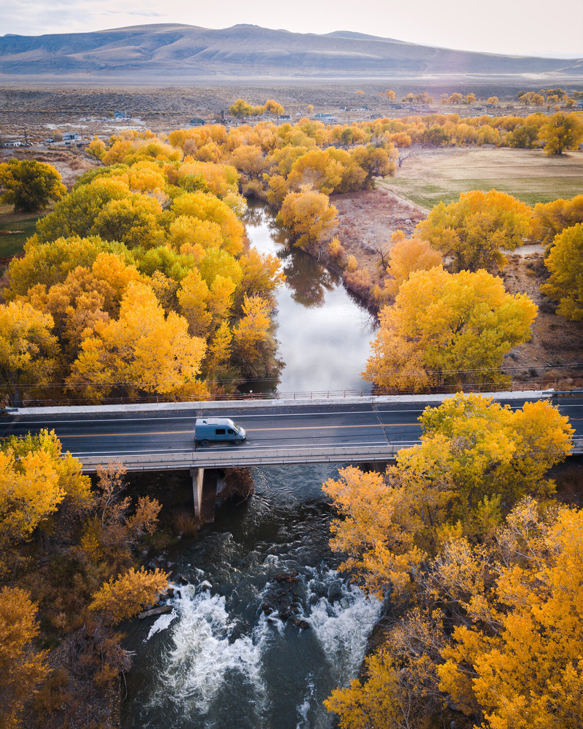 Car driving on a bridge crossing water surrounded by trees with yellow leaves and mountain in the background