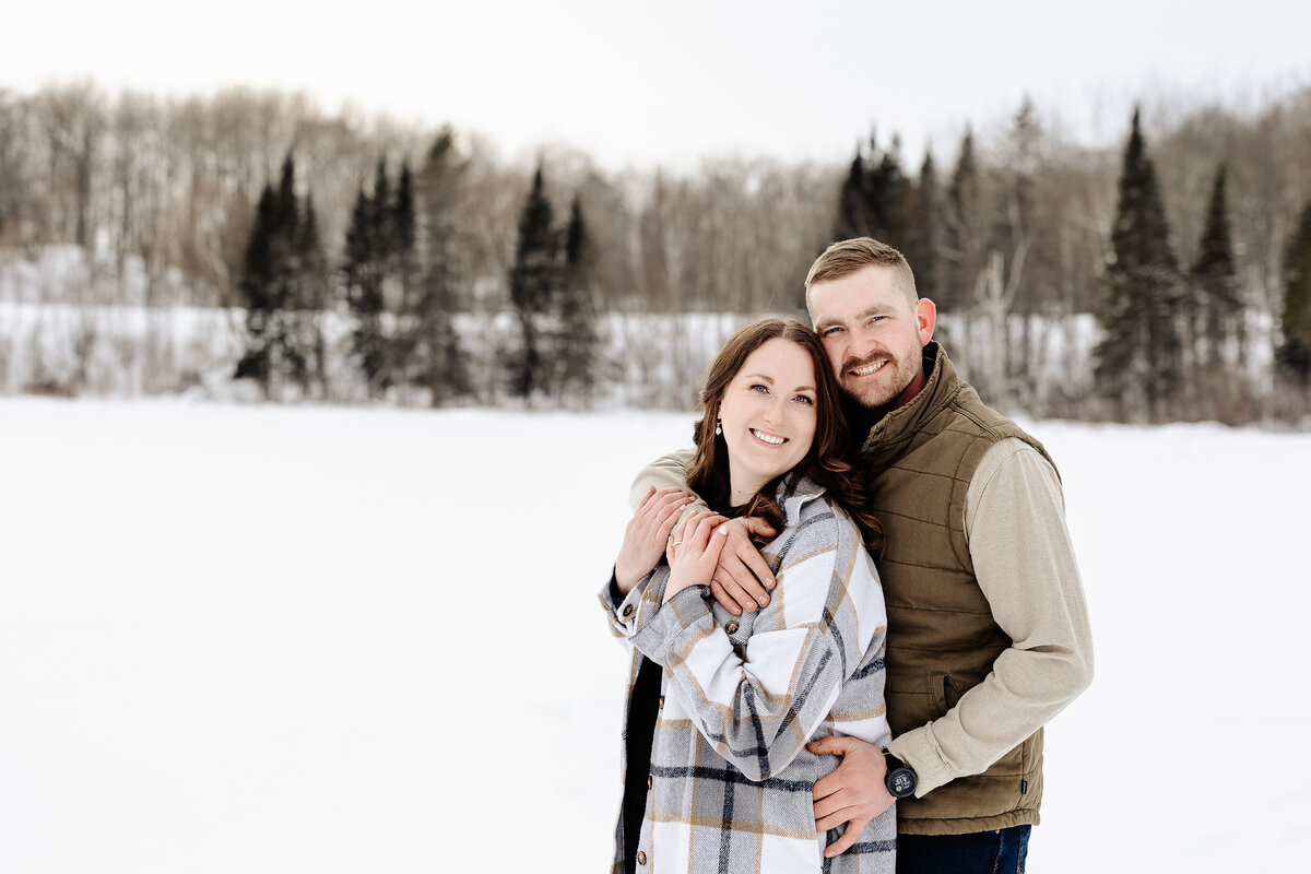 A couple stands in a snowy landscape, smiling warmly. The woman wears a plaid coat, and the man is in a vest. Evergreen trees line the background under a cloudy sky, perfectly capturing the essence of an Amber Bauer Photography family portrait.