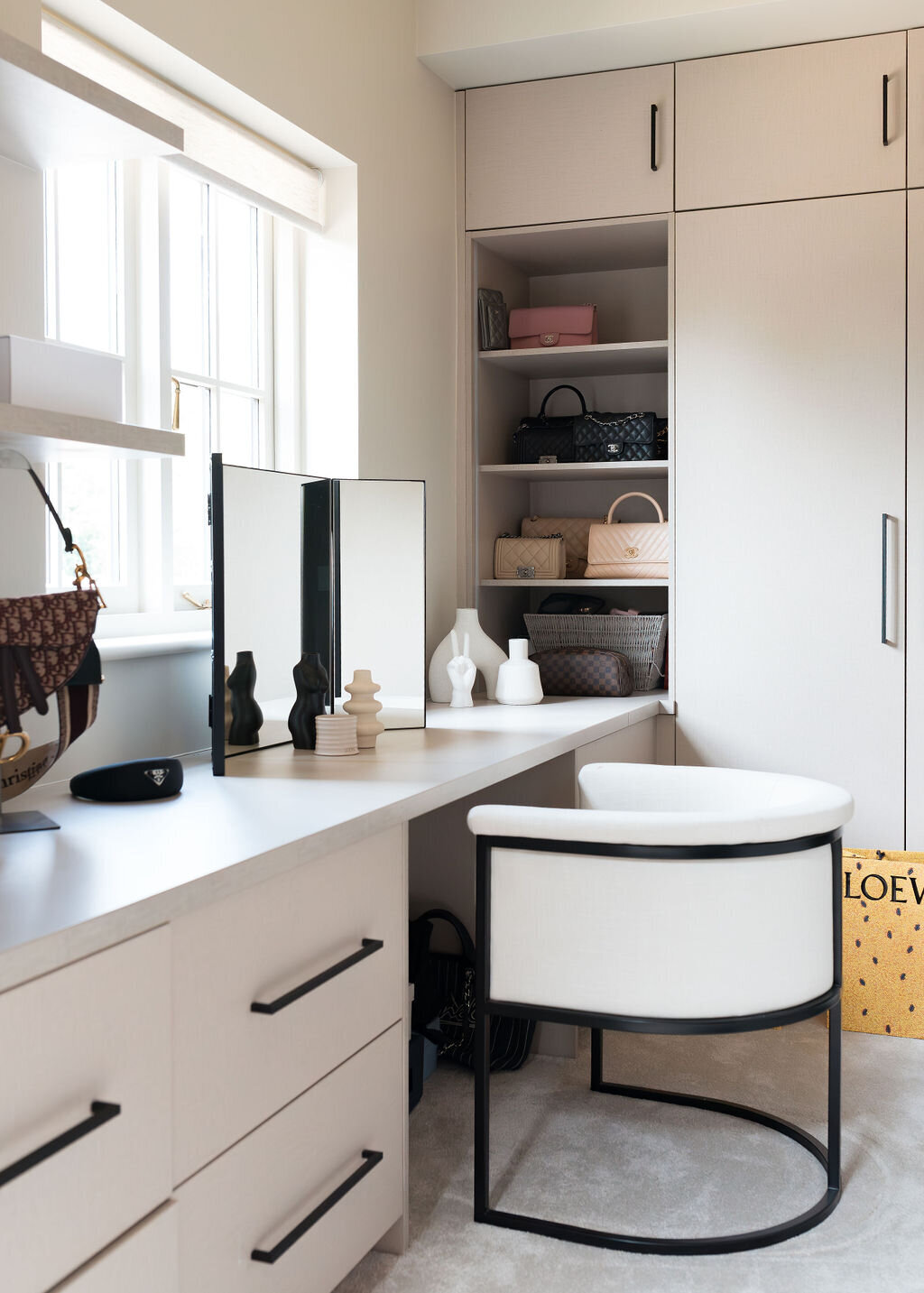 A modern, light-filled walk-in closet with white cabinets and built-in shelves containing handbags and storage baskets. A white desk with a black-framed chair is in the corner, and the floor is carpeted in light neutral.