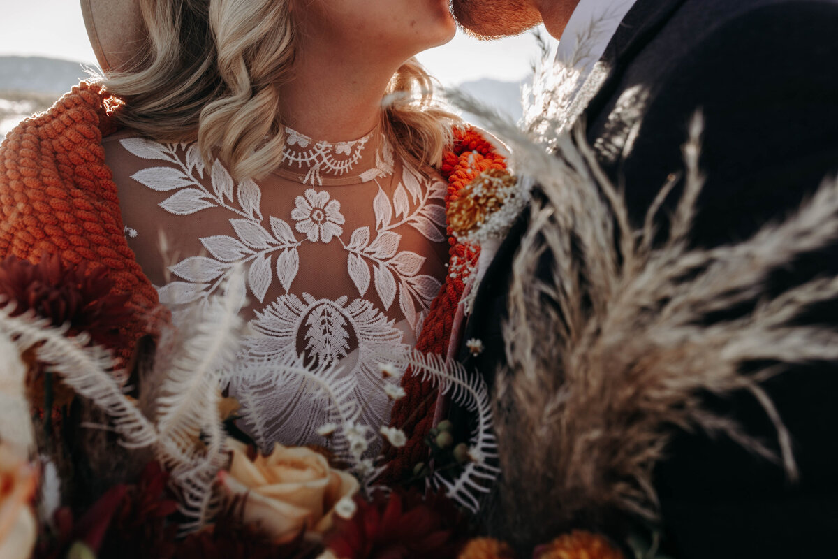 wedding dress with thick lace with a burnt orange blanket wrapped around the bride as she holds a boho floral bouquet and kisses her groom