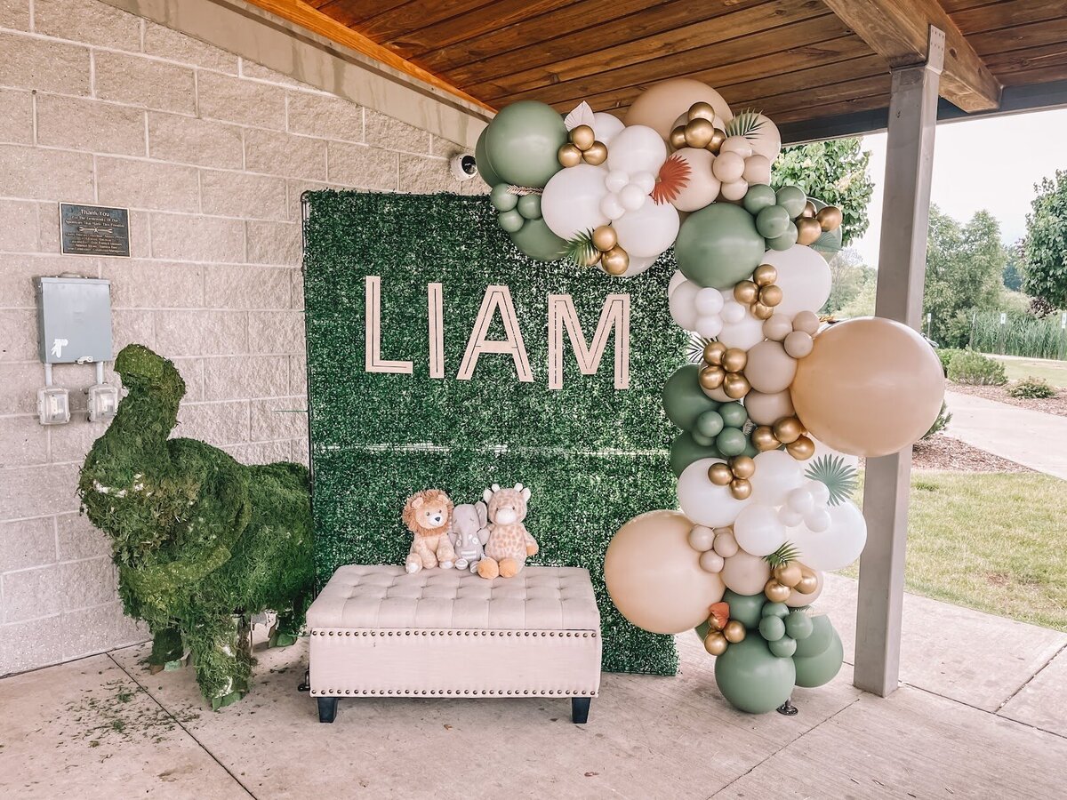 Green backdrop for a baby shower with Liam letters and green, white, and tan balloons.