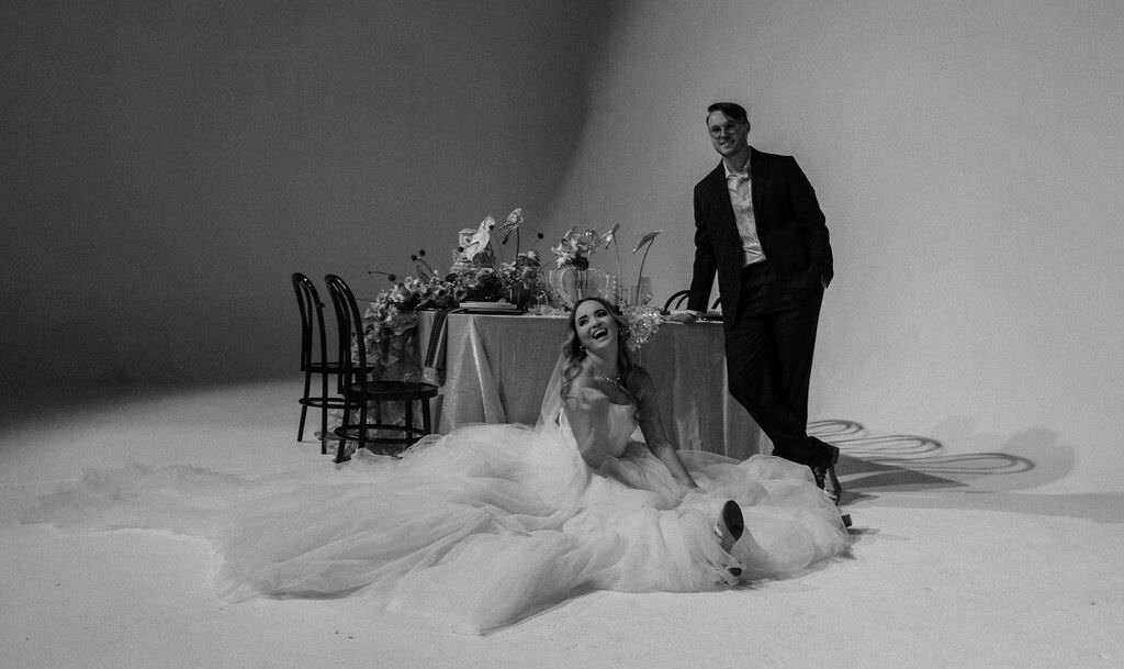 A person on a wedding dress sitting on the ground with their partner standing next to them.